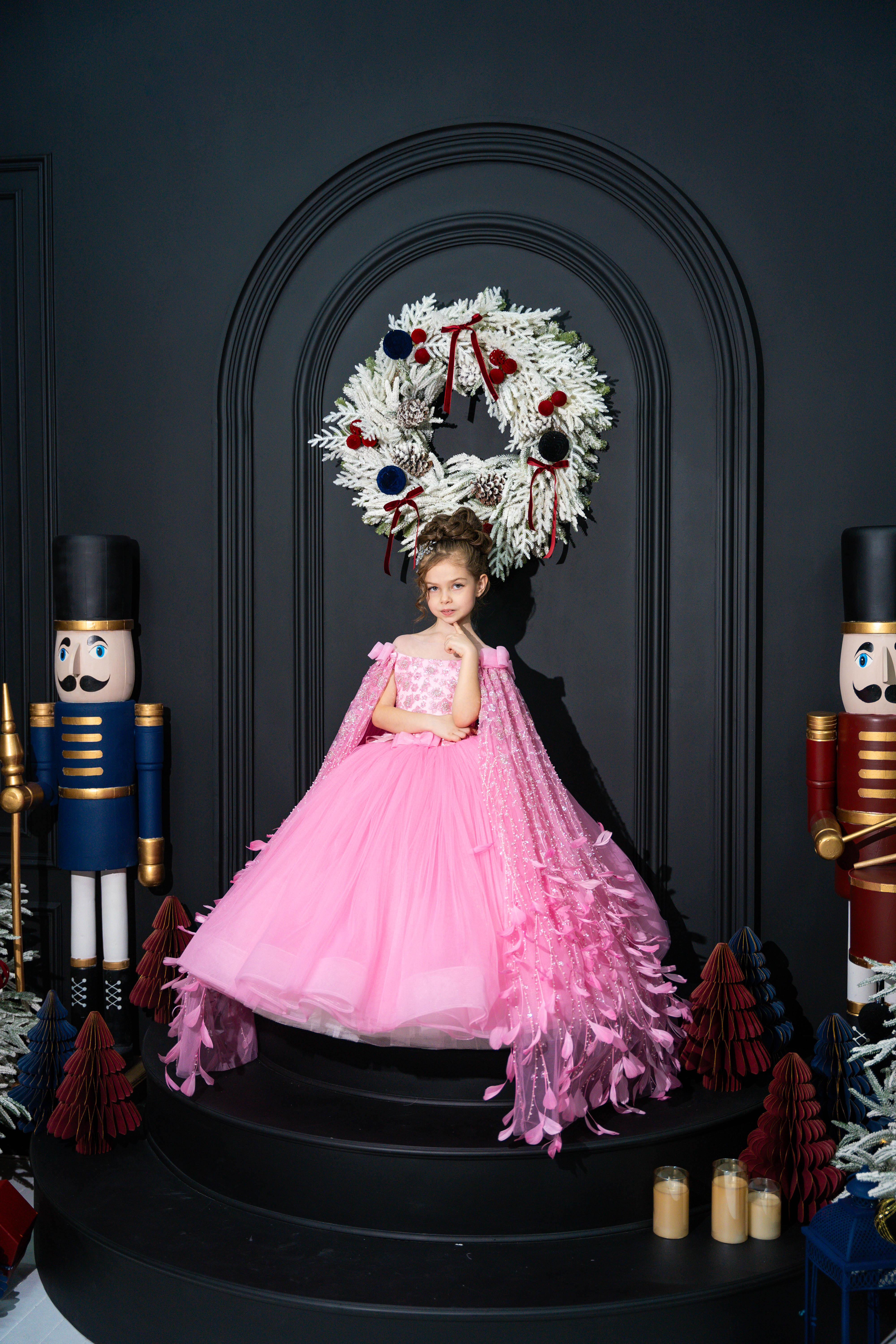 Baby girl in a pink dress standing in front of a decorative wreath with nutcrackers on either side.