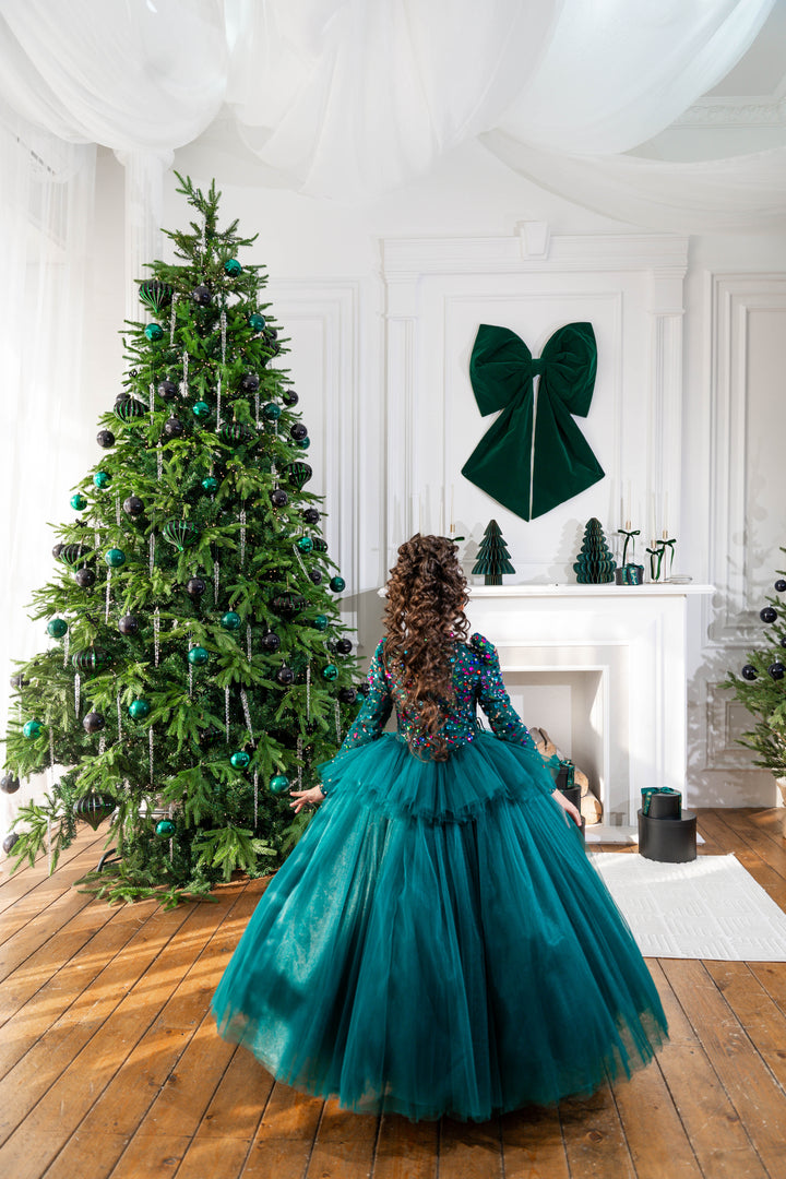 Child in a teal dress standing in front of a decorated Christmas tree in a room with white walls and wooden floors.