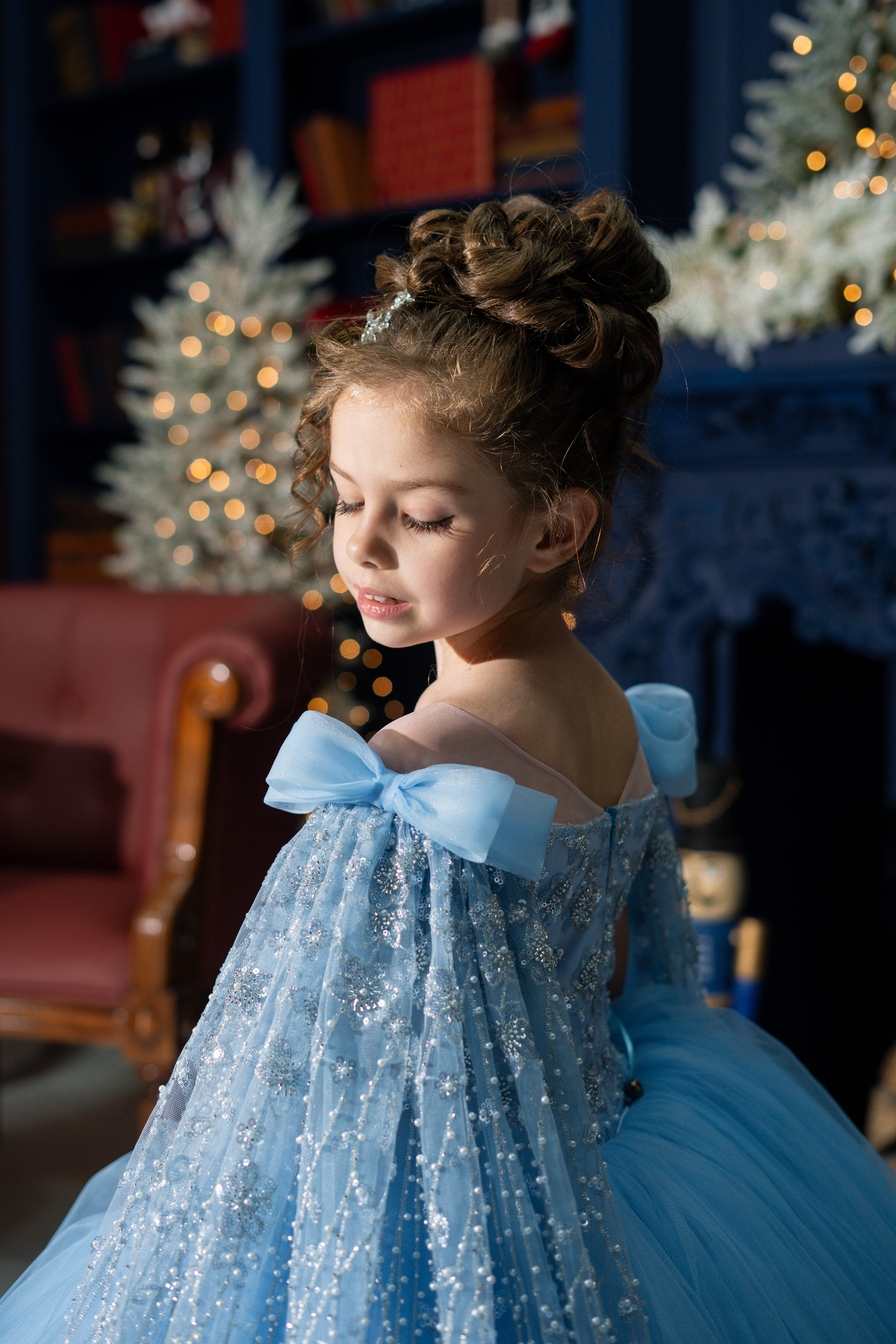 Young girl in a blue dress with a large bow standing in a festive room with Christmas decorations.