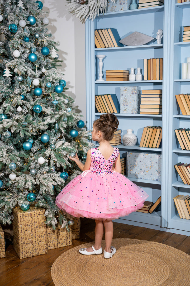 Child in a pink dress standing next to a decorated Christmas tree in a room with bookshelves.