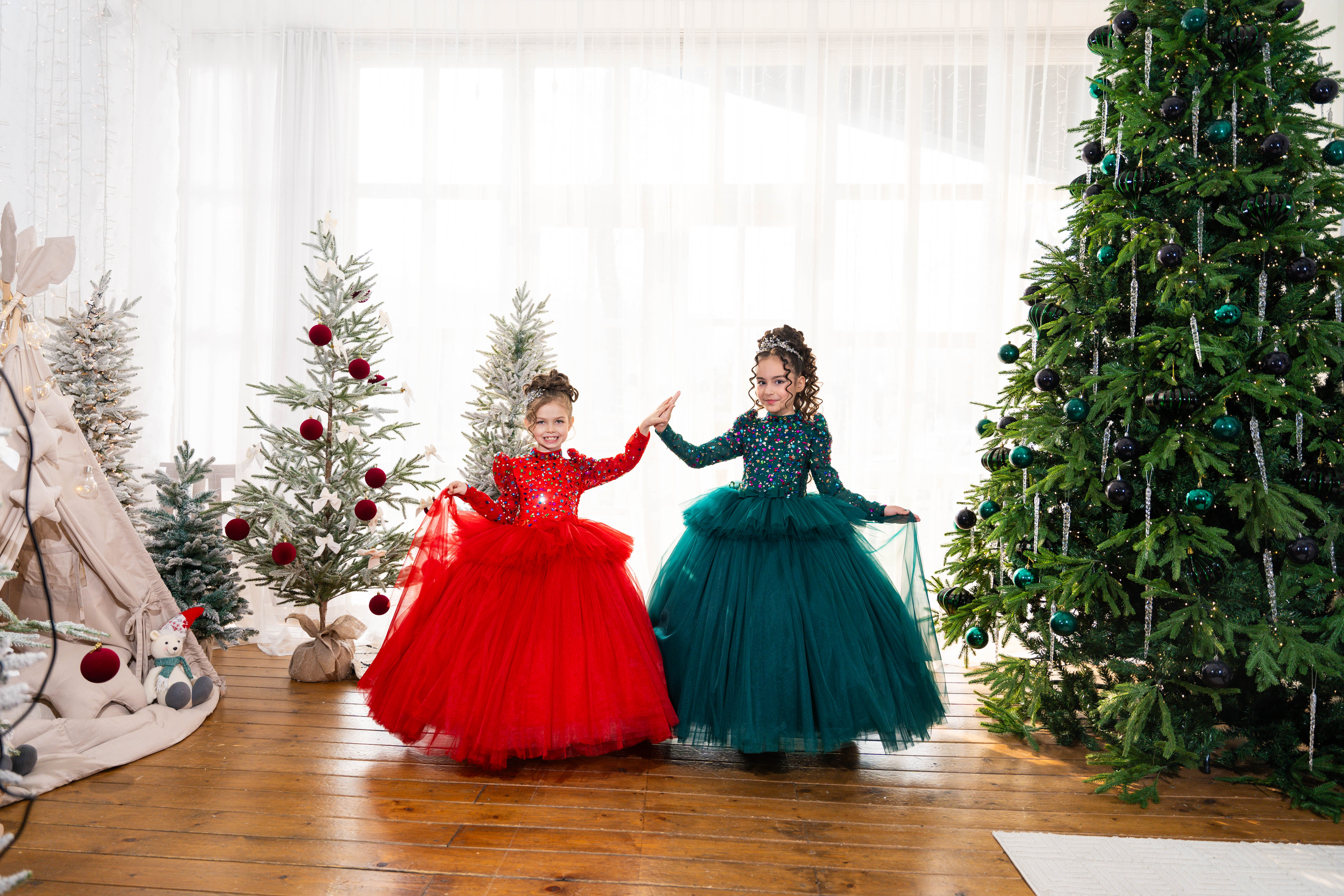 Two children in red and green dresses standing in a room with Christmas trees.