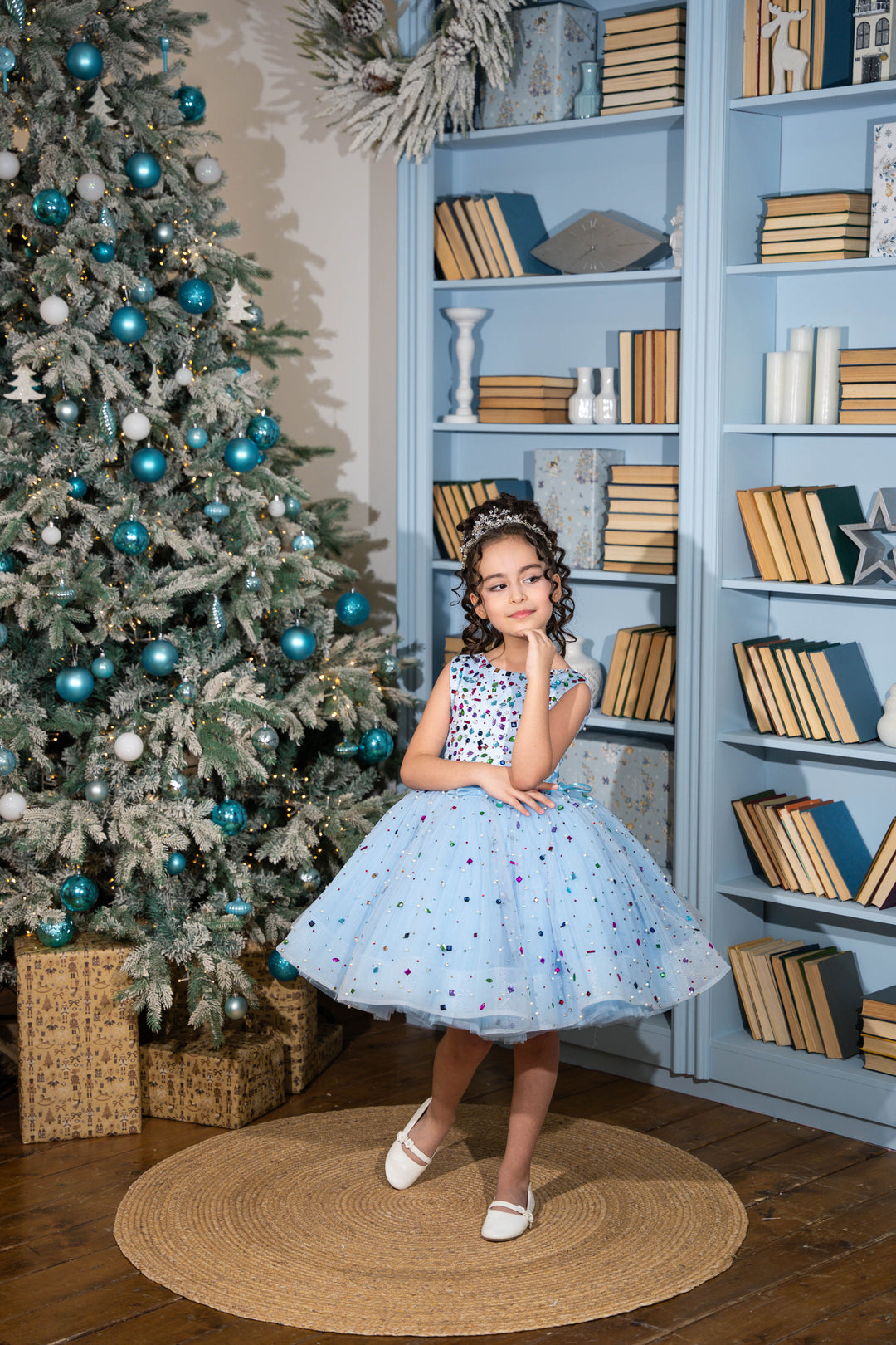 Young girl in a polka dot dress standing in front of a decorated Christmas tree and bookshelf.