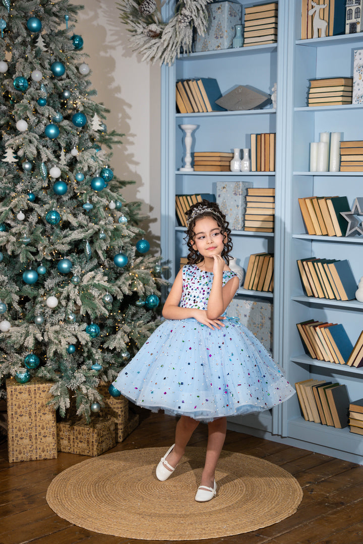 Young girl in a polka dot dress standing in front of a decorated Christmas tree and bookshelf.