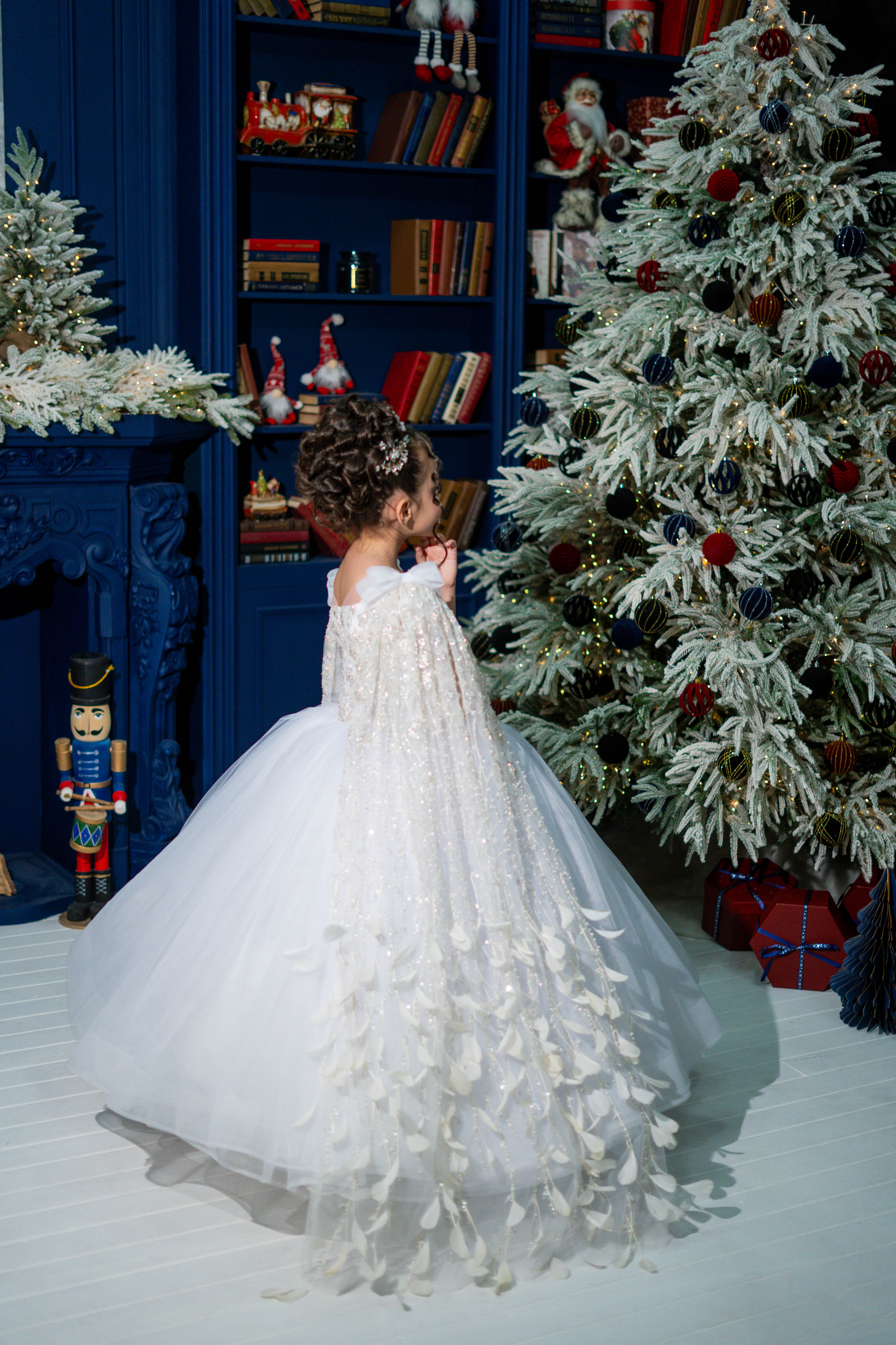 Child in a white dress standing in front of a decorated Christmas tree and bookshelf.