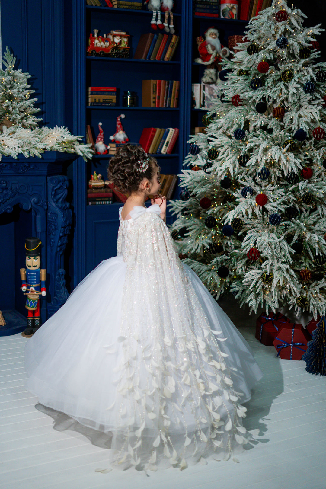 Child in a white dress standing in front of a decorated Christmas tree and bookshelf.