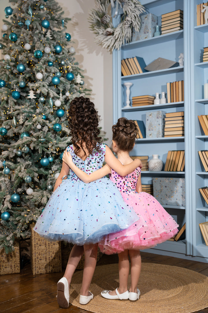 Two children in colorful dresses standing in front of a decorated Christmas tree and bookshelf.