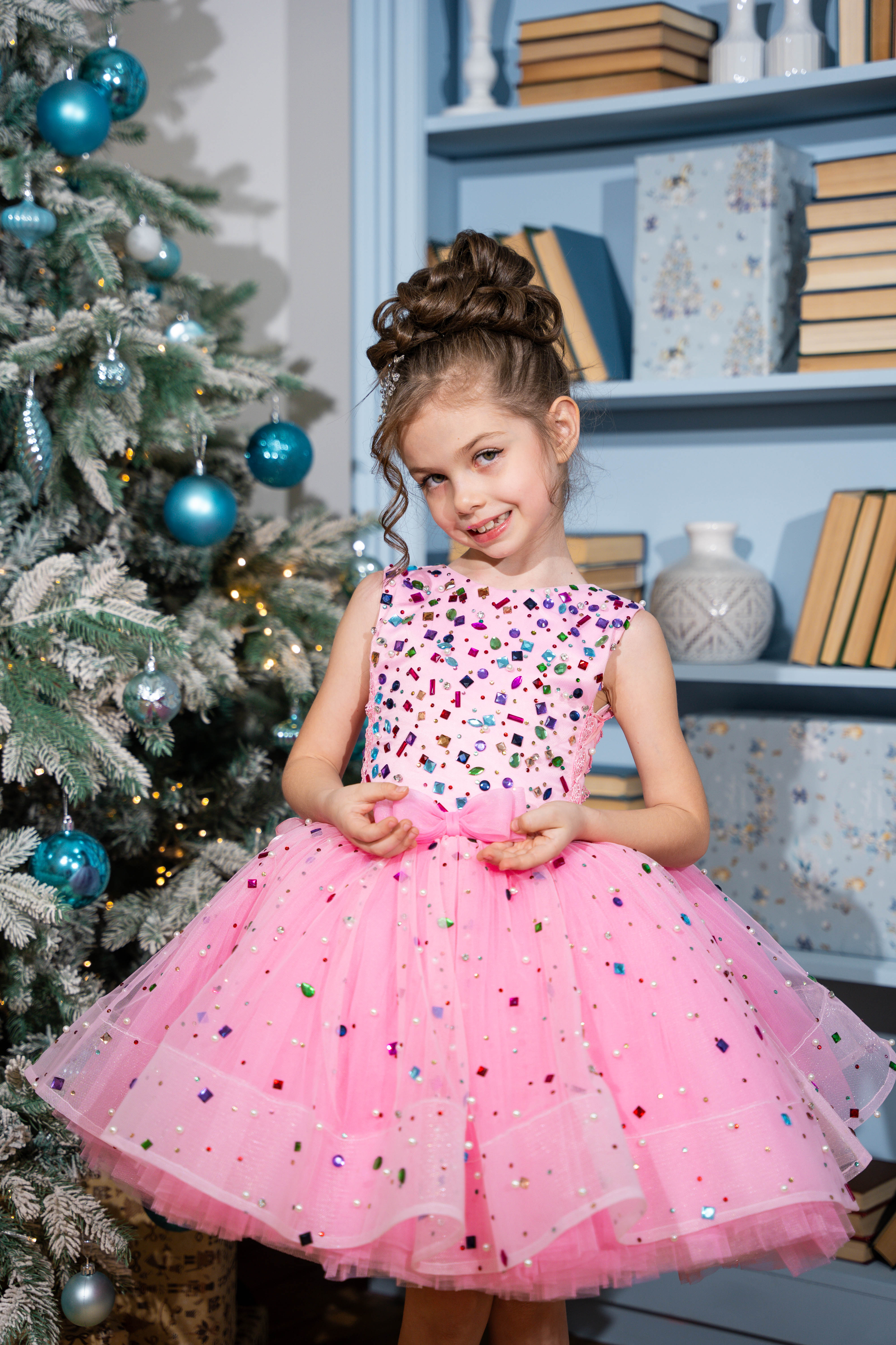Baby girl in a pink dress with colorful polka dots standing next to a decorated Christmas tree.