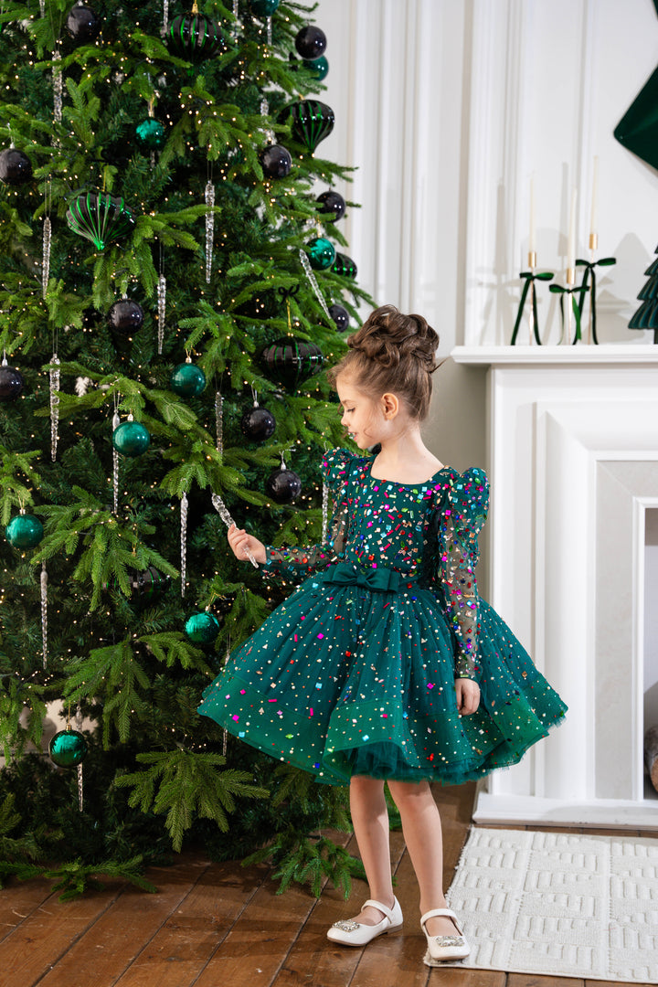 Baby girl in a green dress standing next to a decorated Christmas tree.