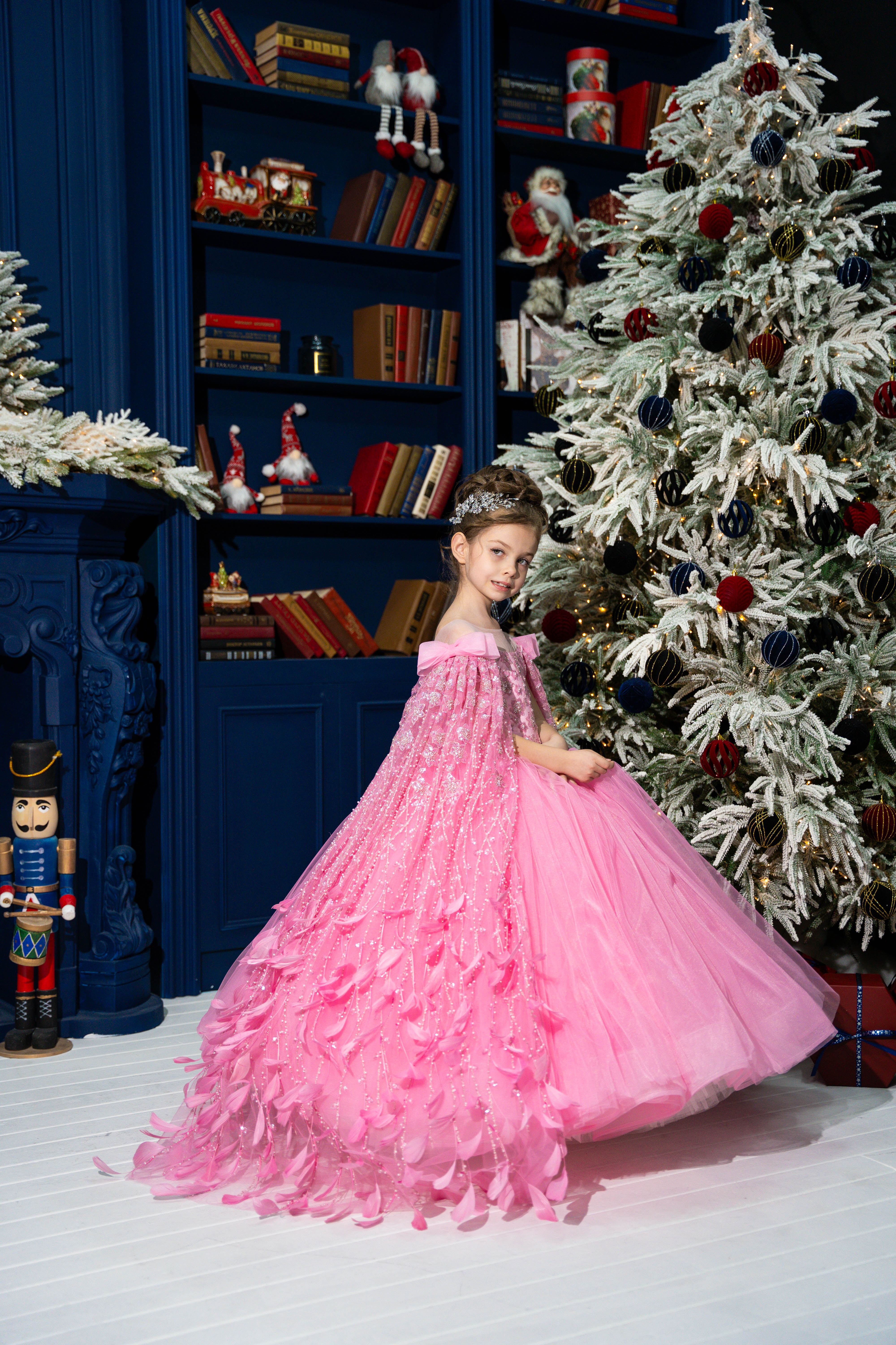 Child in a pink dress standing in front of a decorated Christmas tree and bookshelf.