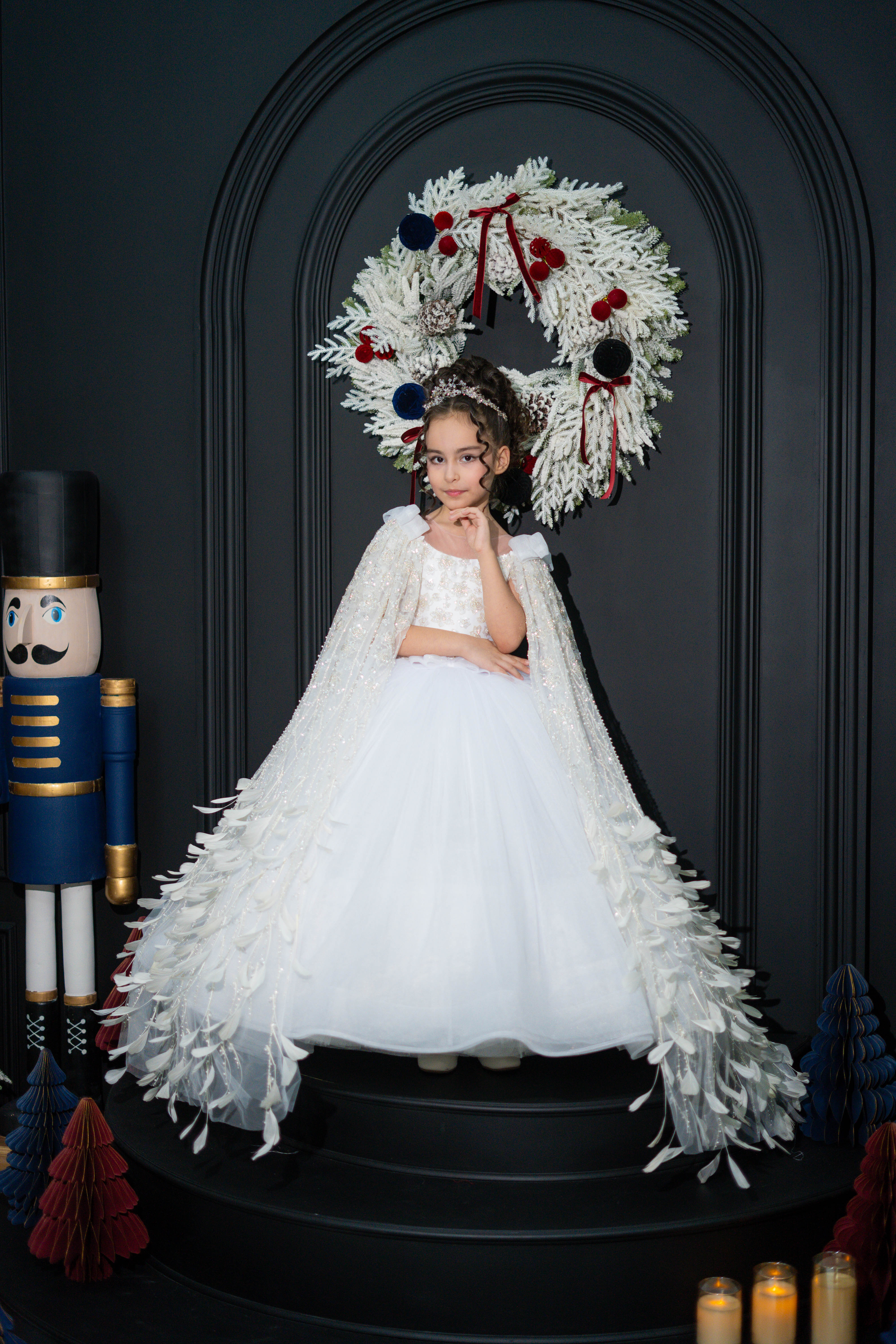A child in a white dress standing in front of a decorative wreath on a dark background