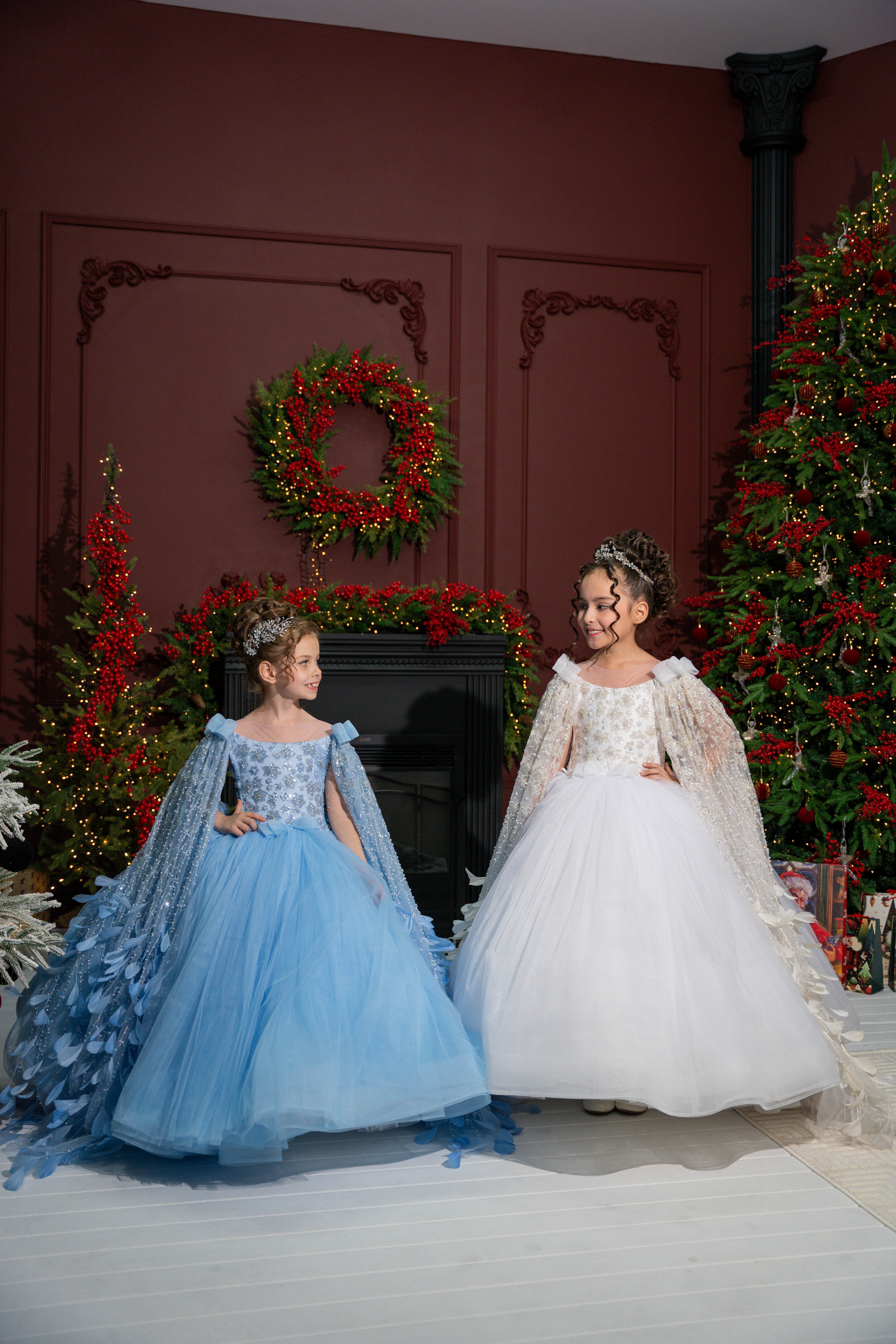 Two children in festive costumes standing in a decorated room with Christmas trees and wreaths.