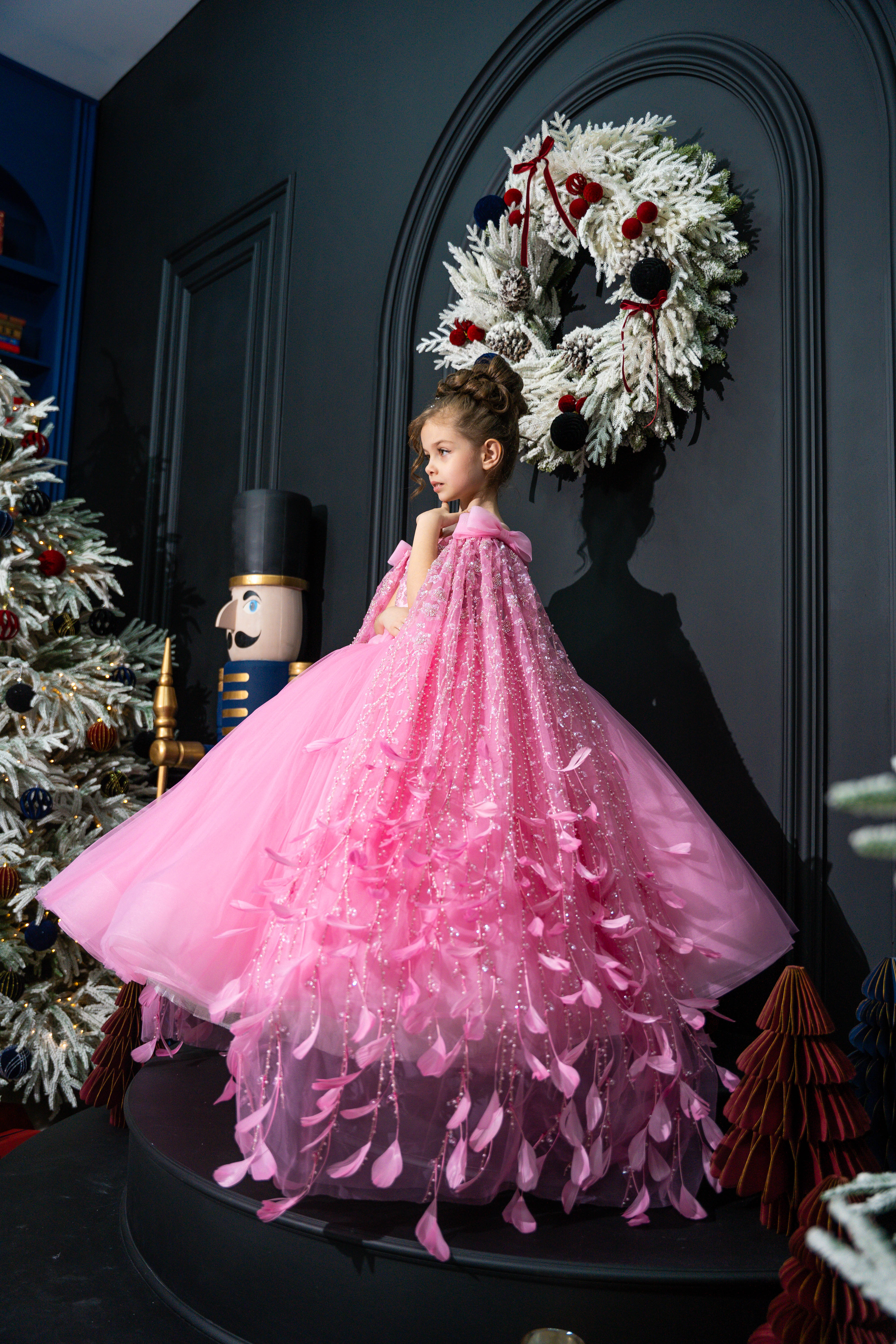 Child in a pink dress standing in front of a decorated wall with a wreath.
