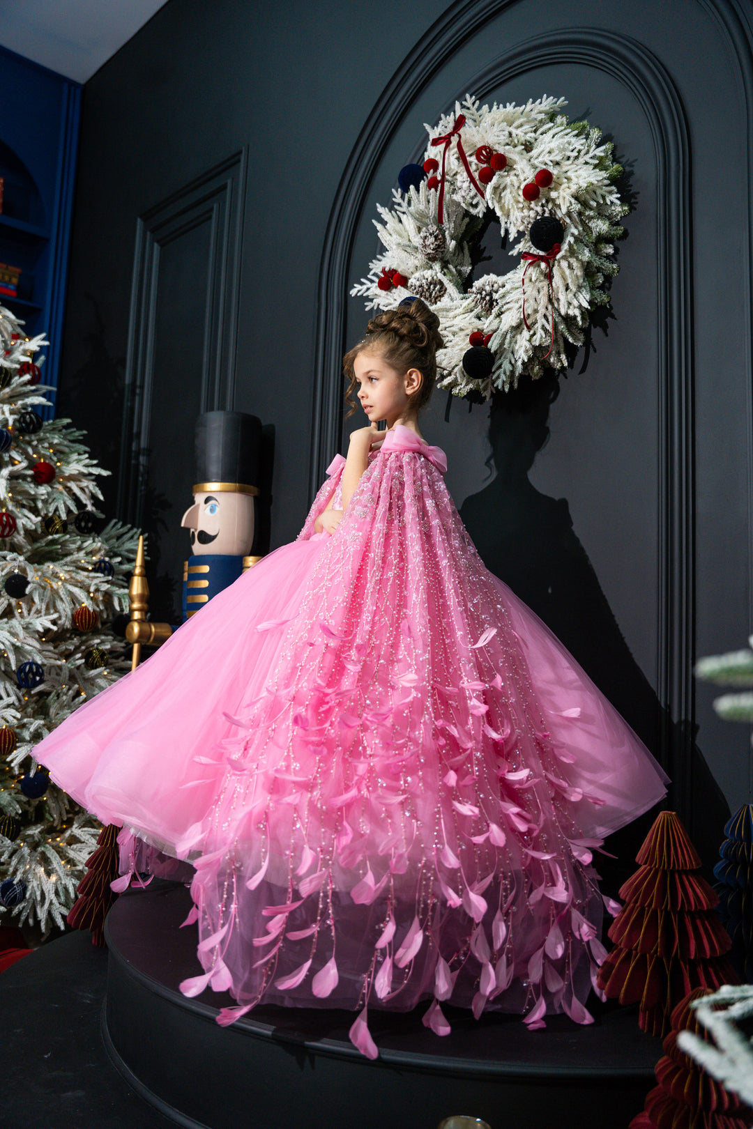 Child in a pink dress standing in front of a decorated wall with a wreath.