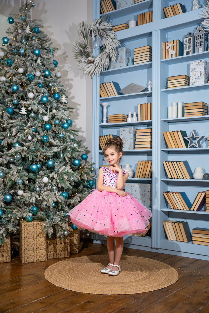 Baby girl in a pink dress standing in front of a decorated Christmas tree and bookshelf.