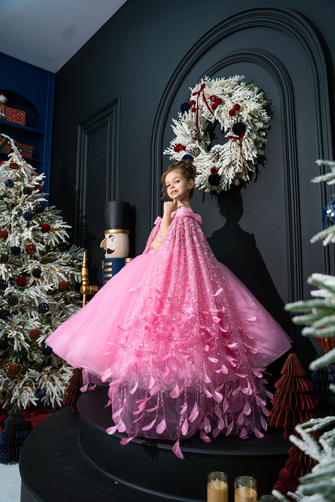 Child in a pink dress standing in front of a decorated Christmas wreath.
