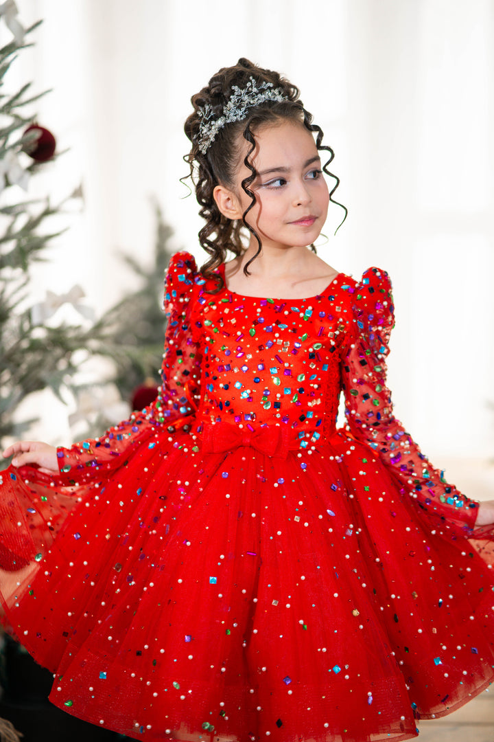 Baby girl in a red dress with sequins standing in front of a blurred Christmas tree.