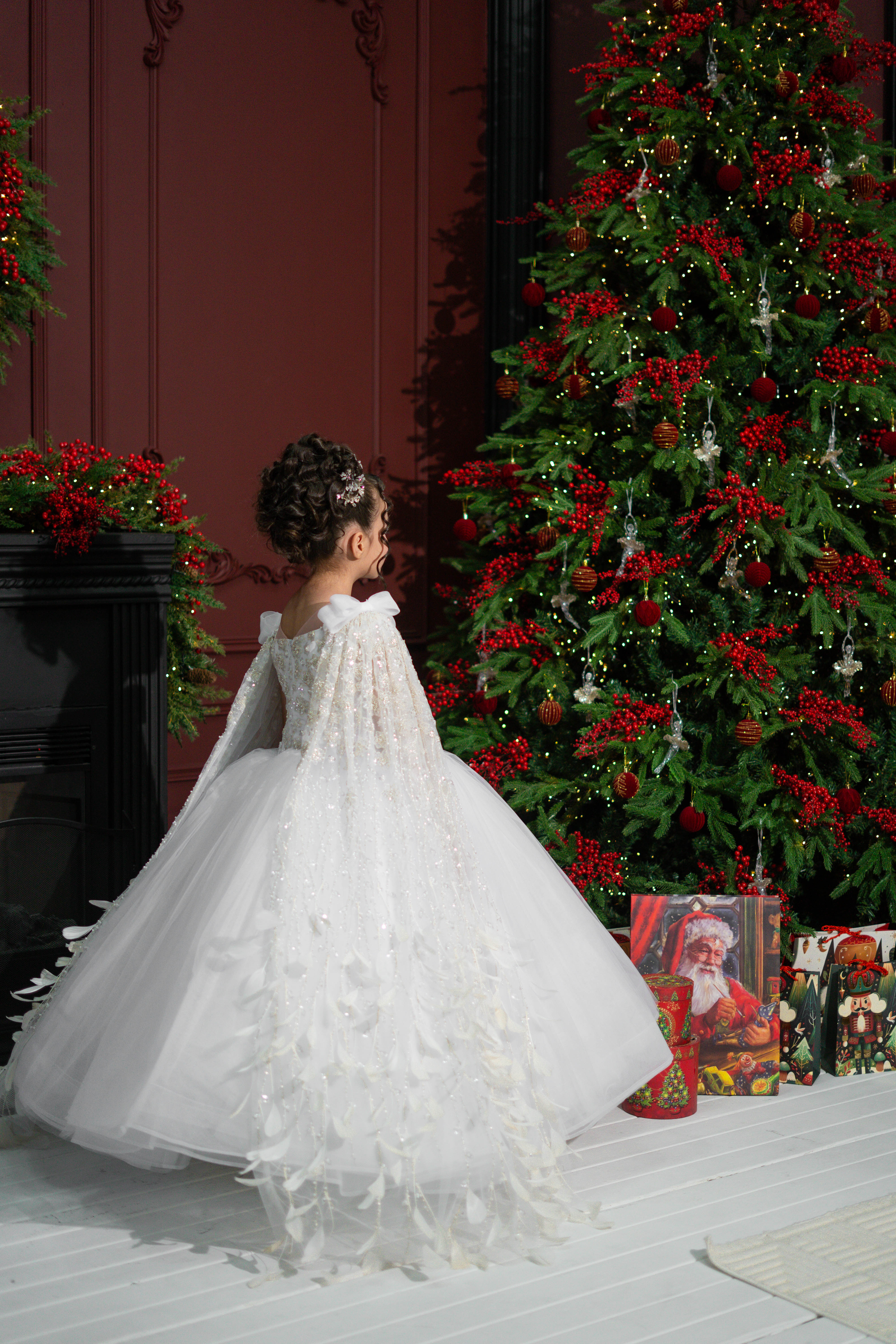 A child in a white dress standing in front of a decorated Christmas tree with red berries.