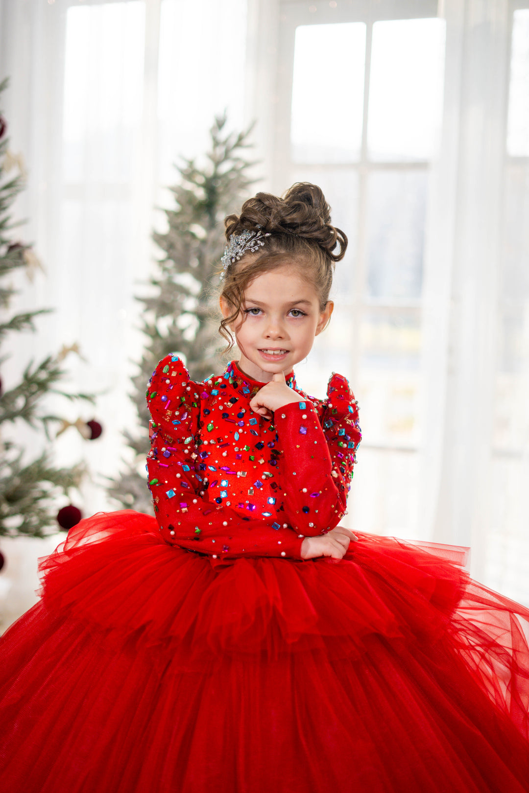 Baby girl in a red dress with sequins and a tulle skirt, standing in front of a decorated Christmas tree.