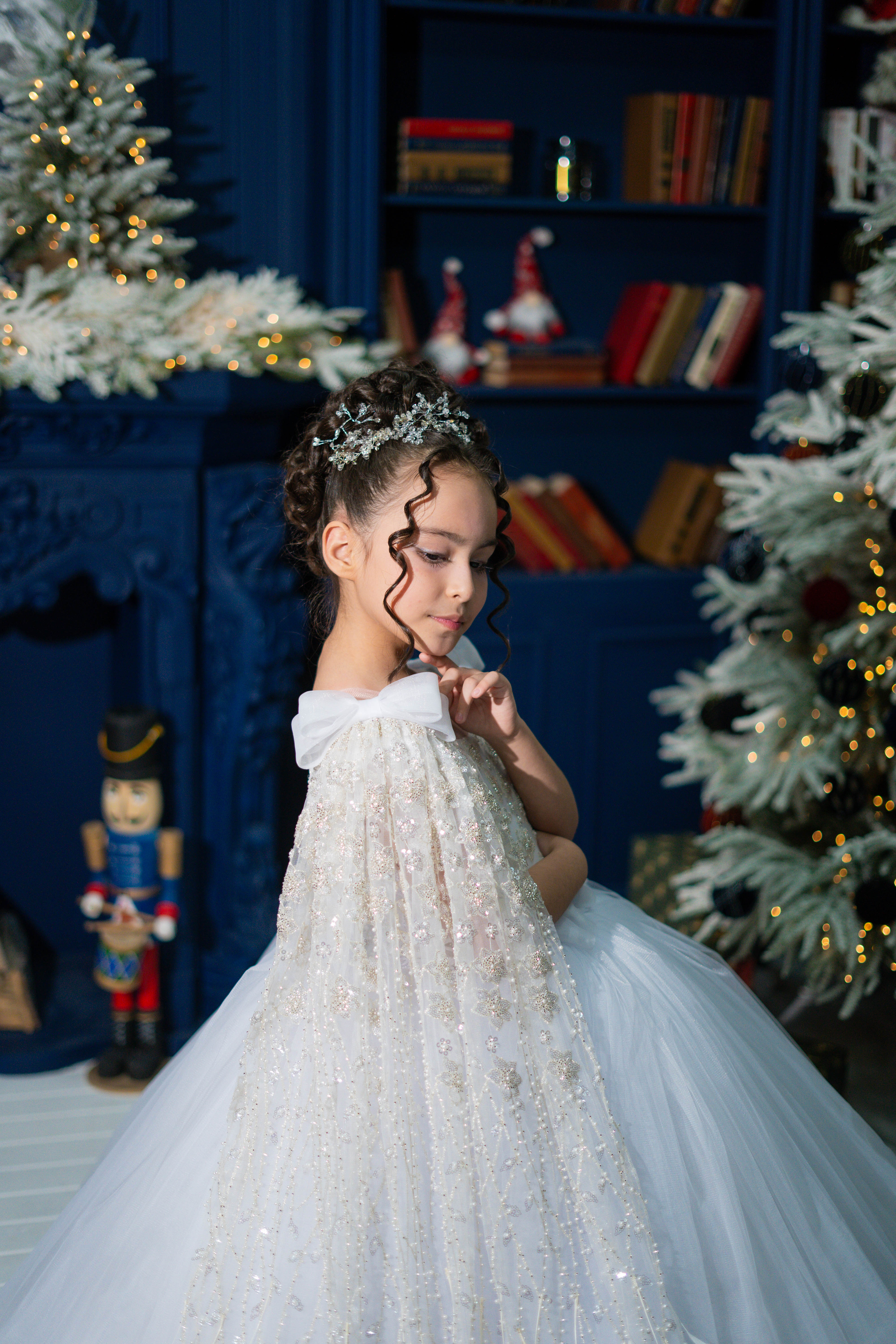 Baby girl in a white dress standing in front of a decorated Christmas fireplace.