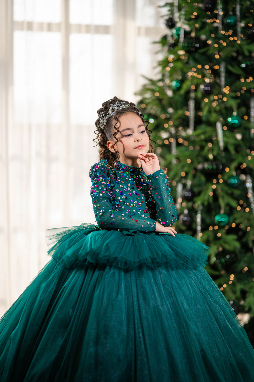 Young girl in a green dress with a tiara, standing in front of a decorated Christmas tree.