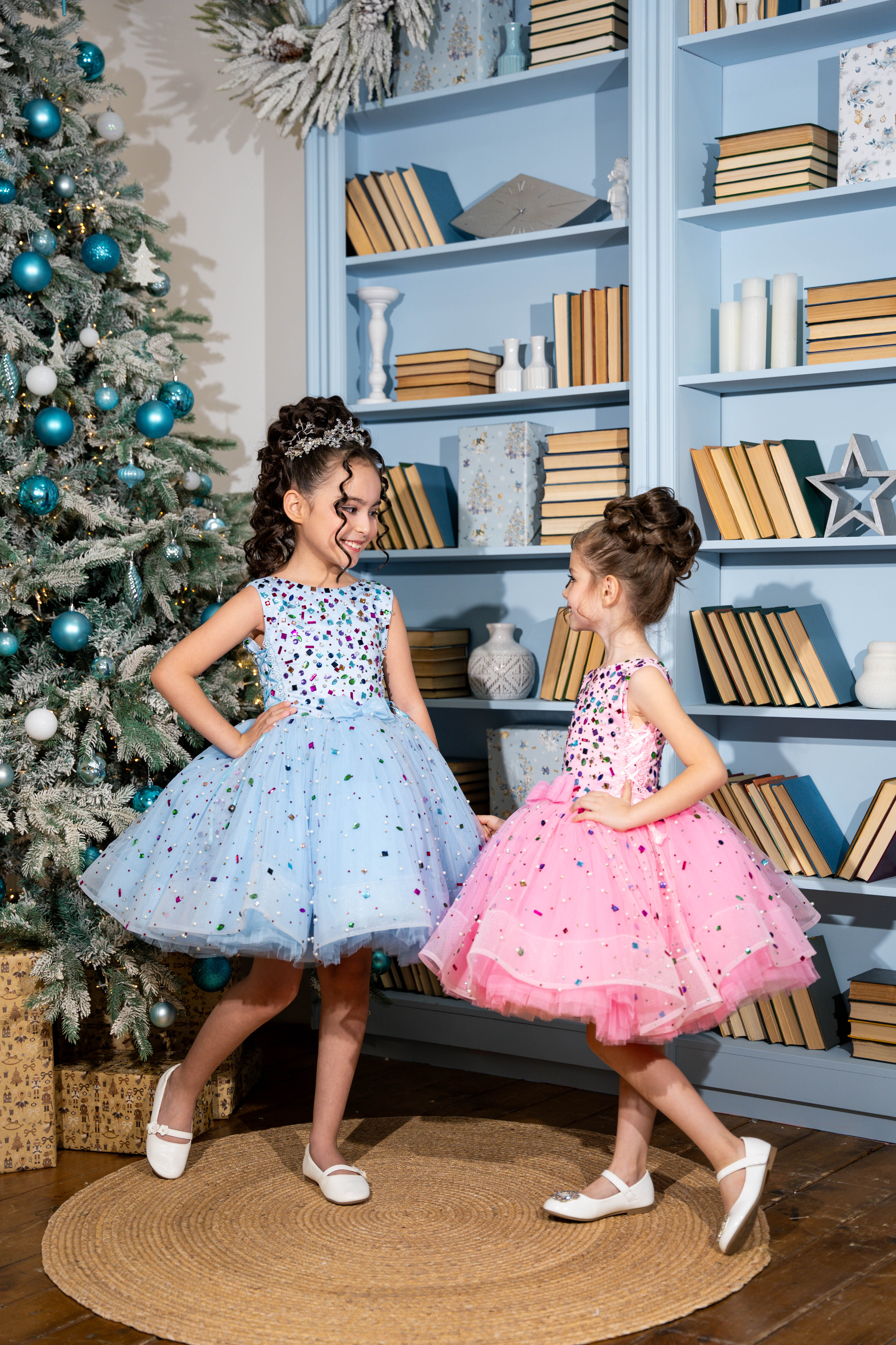 Two young girls in dresses standing in a room with a decorated Christmas tree and bookshelves.