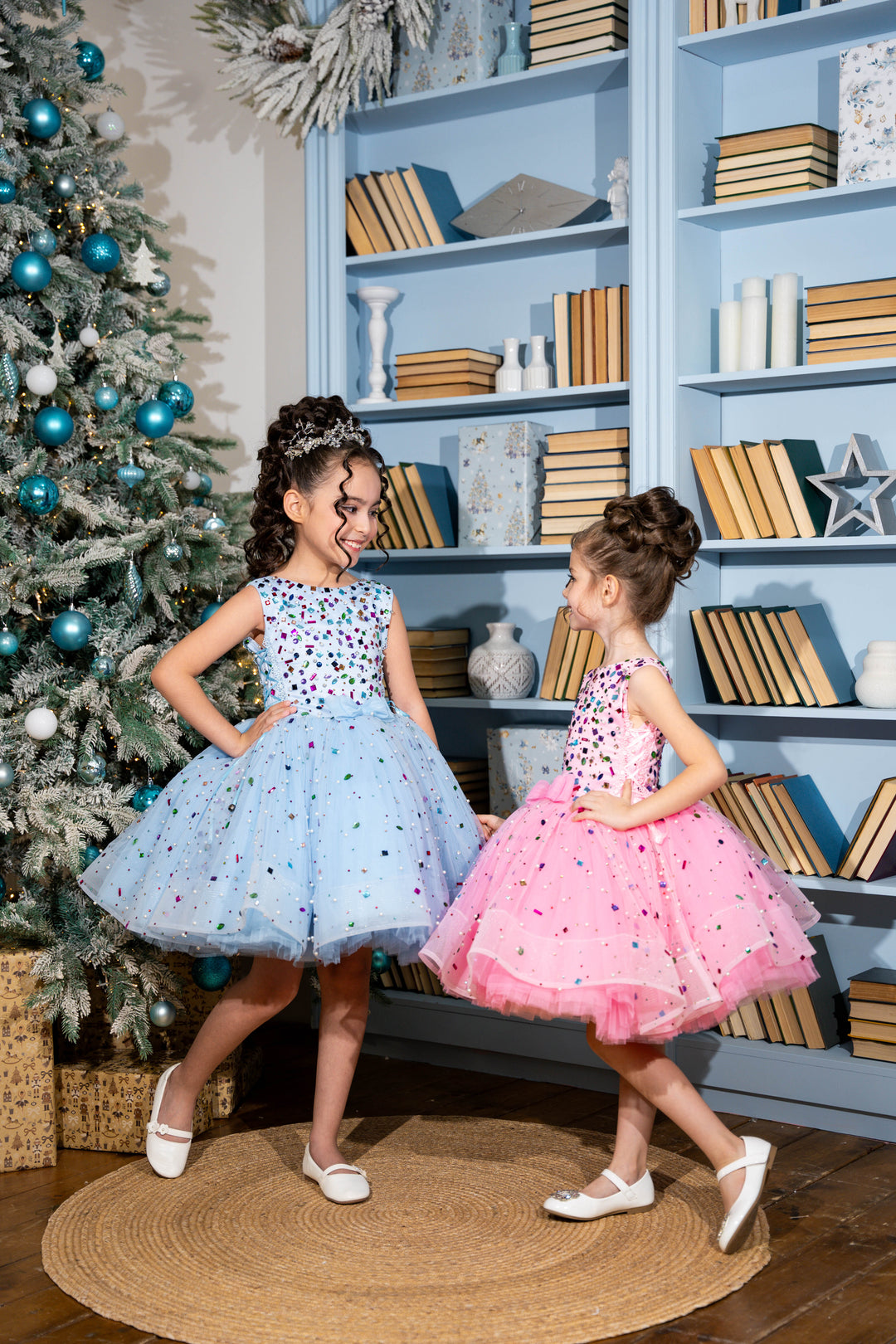 Two young girls in dresses standing in a room with a decorated Christmas tree and bookshelves.