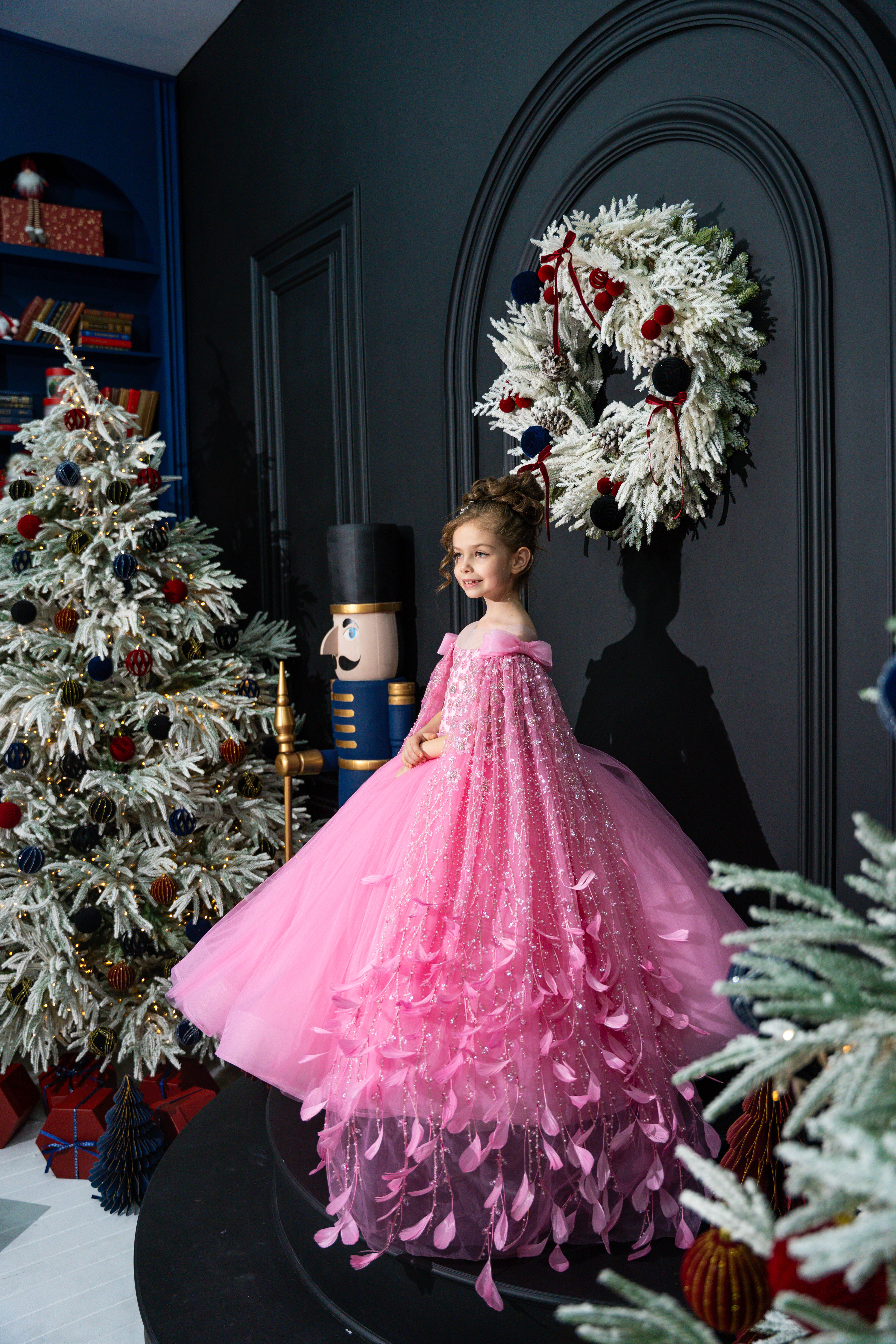 Child in a pink dress standing in a decorated room with Christmas trees and wreaths.
