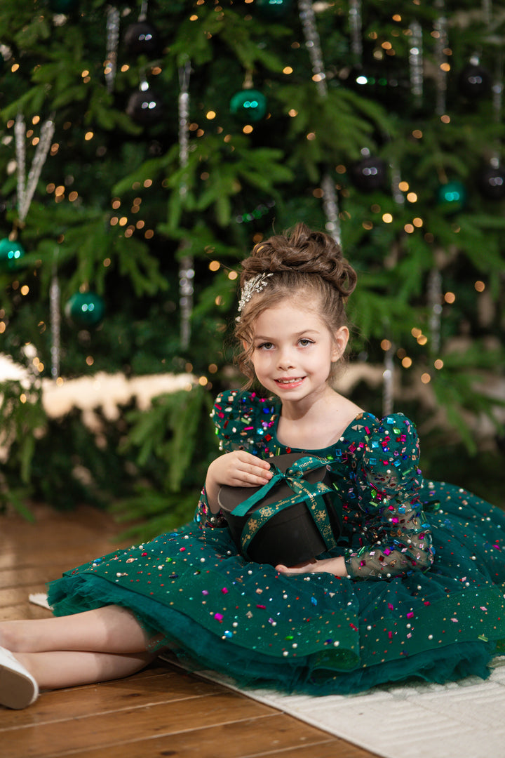 Baby girl in a green dress sitting on the floor in front of a decorated Christmas tree.