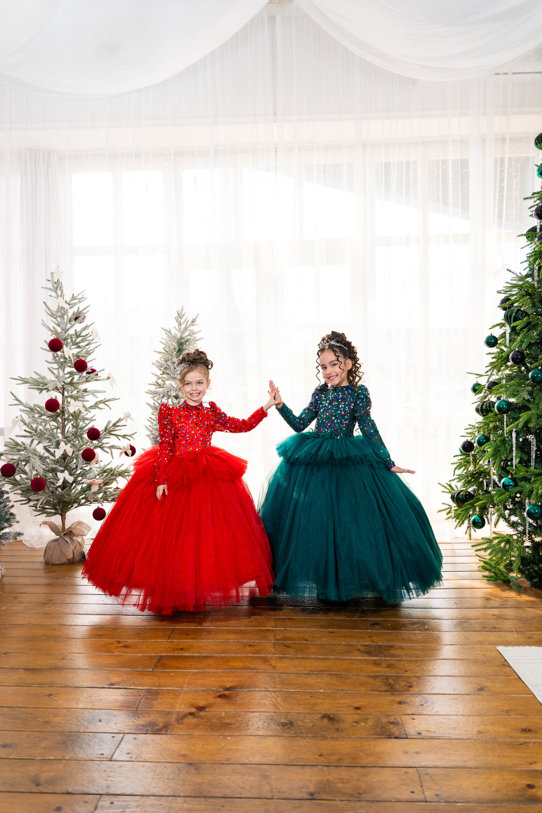 Two children in red and green dresses standing in a room with Christmas trees.