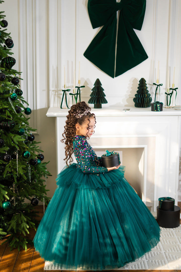 Young girl in a teal dress holding a gift box in front of a decorated Christmas tree and fireplace.