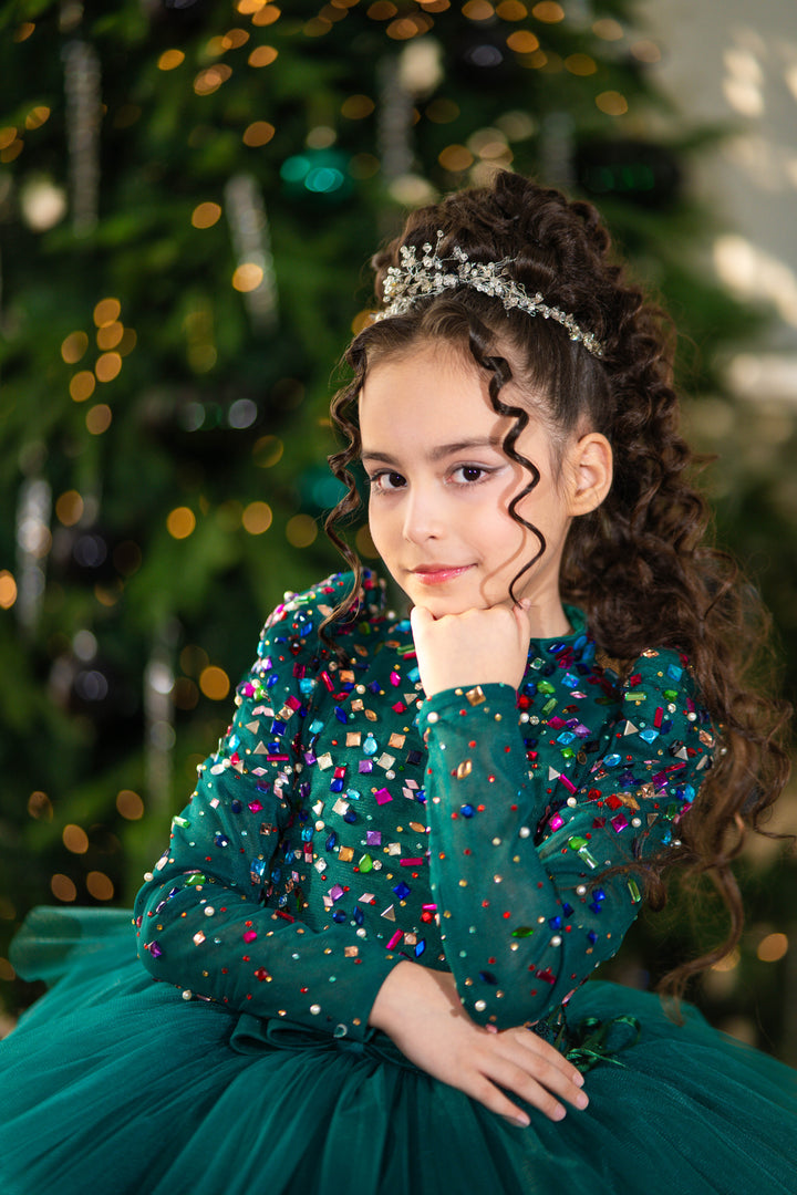 Young girl in a teal dress with colorful sequins and a tiara, standing in front of a decorated Christmas tree.