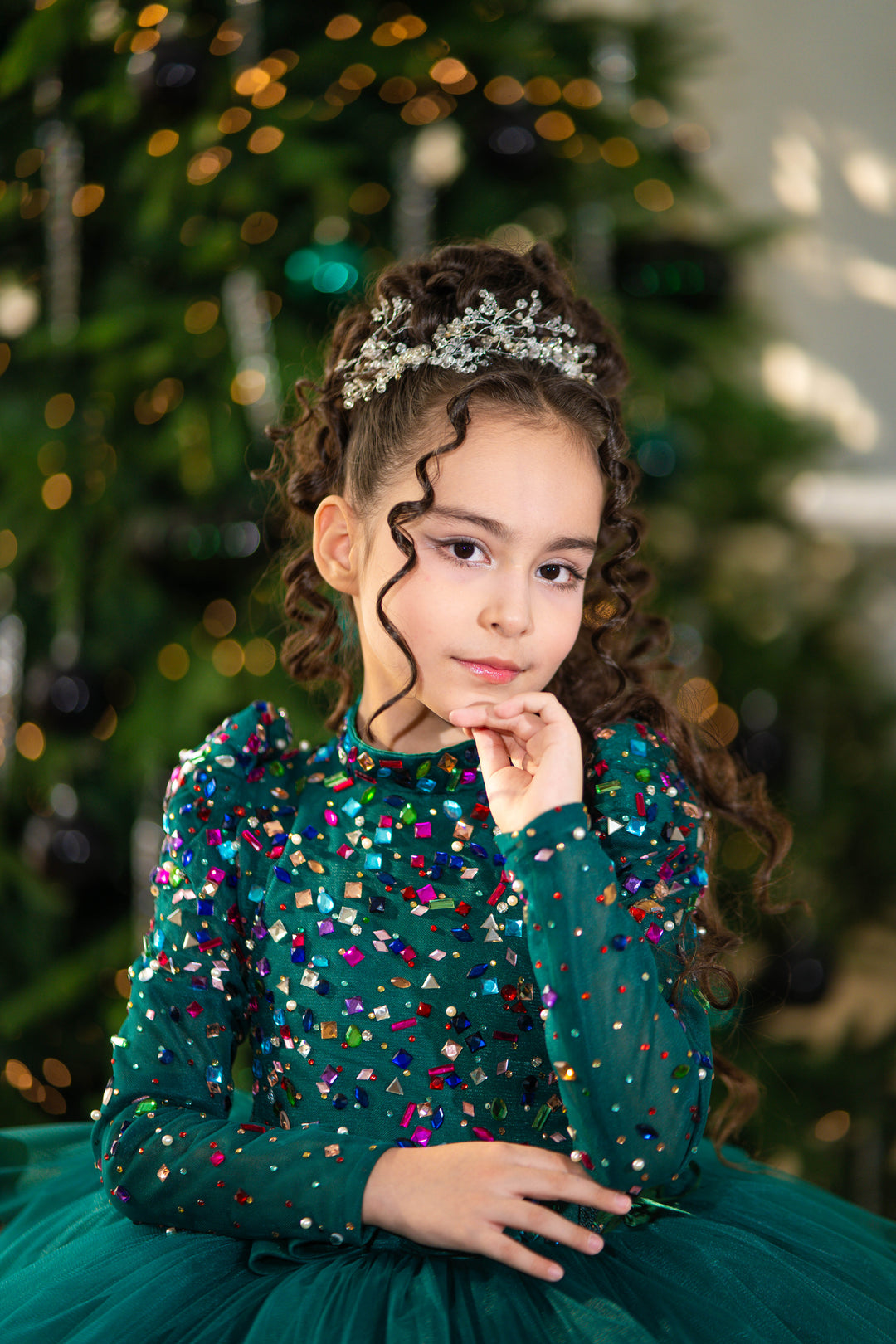 Young girl in a green dress with colorful sequins and a tiara, standing in front of a decorated Christmas tree.