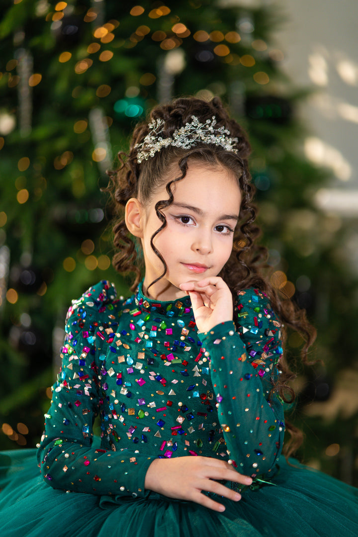 Young girl in a green dress with colorful sequins and a tiara, standing in front of a decorated Christmas tree.