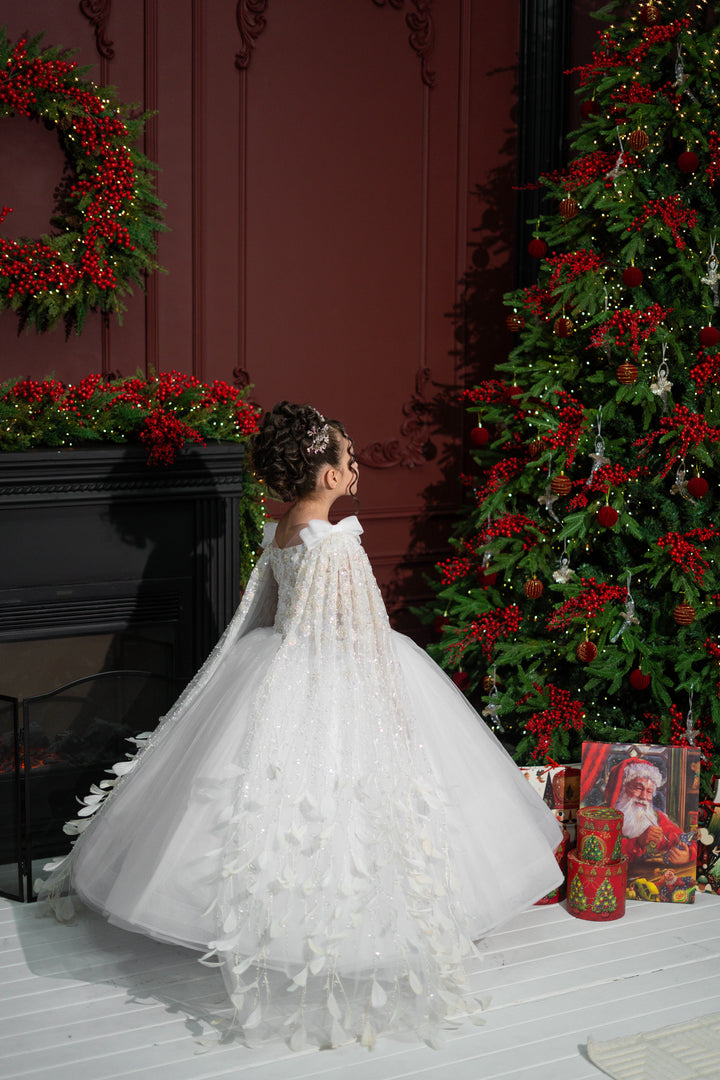 A child in a white dress standing in front of a decorated Christmas tree and fireplace.