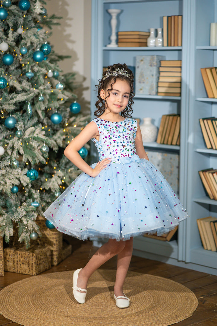 Young girl in a light blue dress with colorful polka dots standing in front of a decorated Christmas tree and bookshelf.