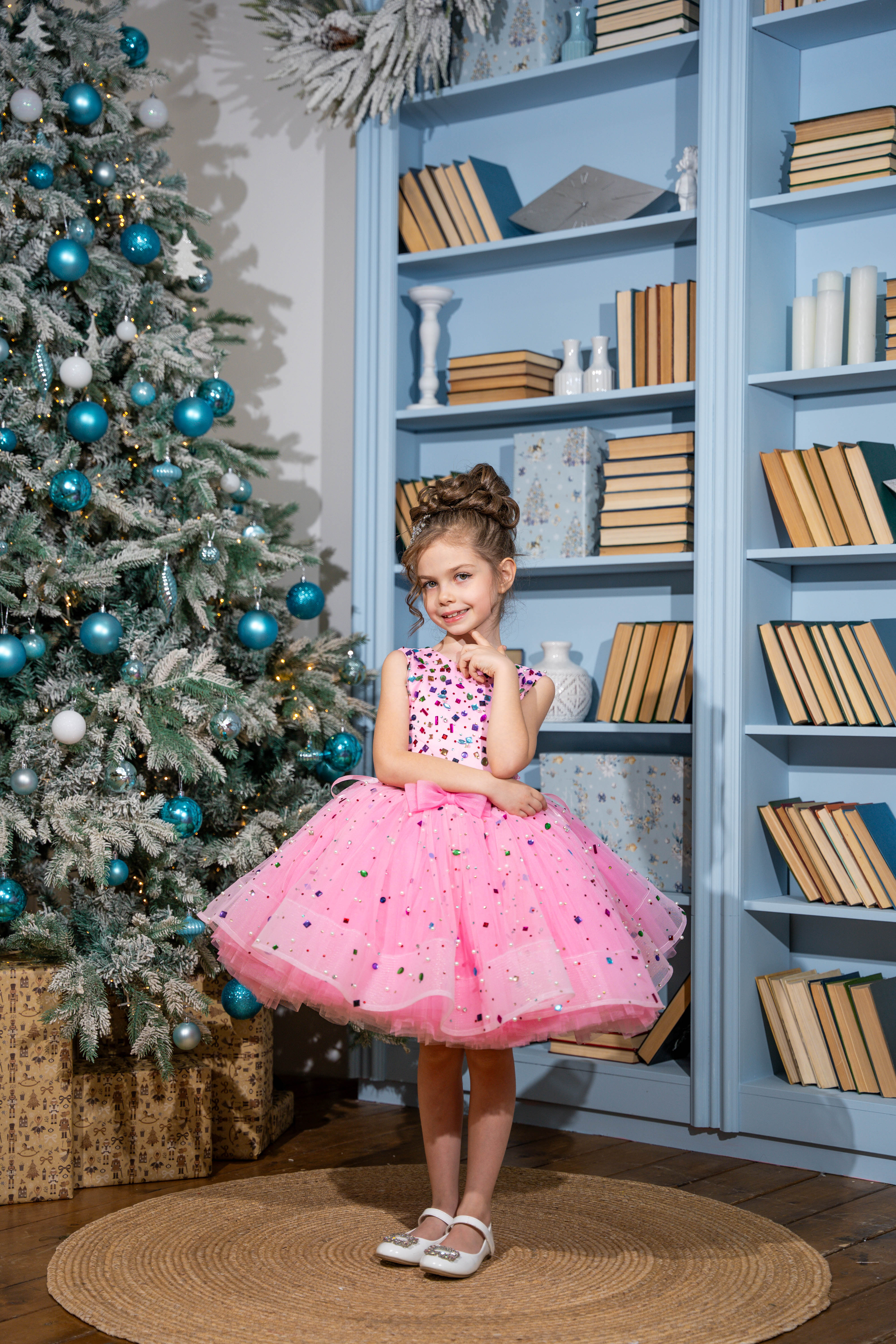 Baby girl in a pink dress standing in front of a decorated Christmas tree and bookshelf.