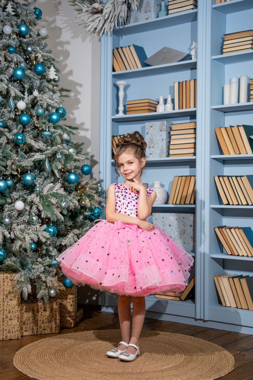 Baby girl in a pink dress standing in front of a decorated Christmas tree and bookshelf.
