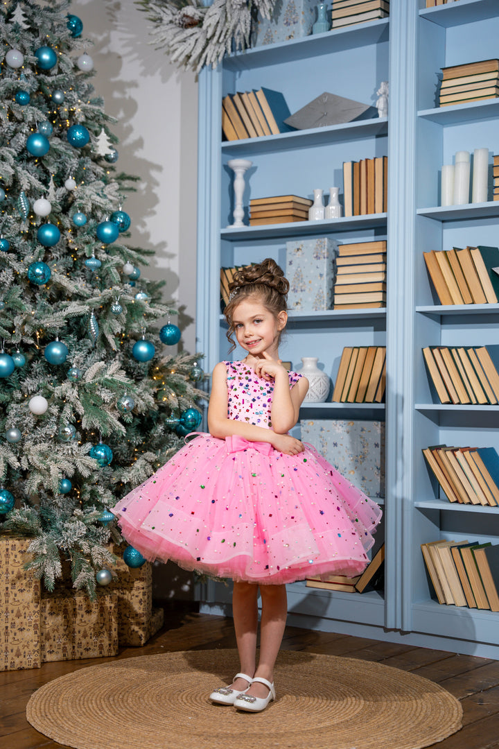 Baby girl in a pink dress standing in front of a decorated Christmas tree and bookshelf.