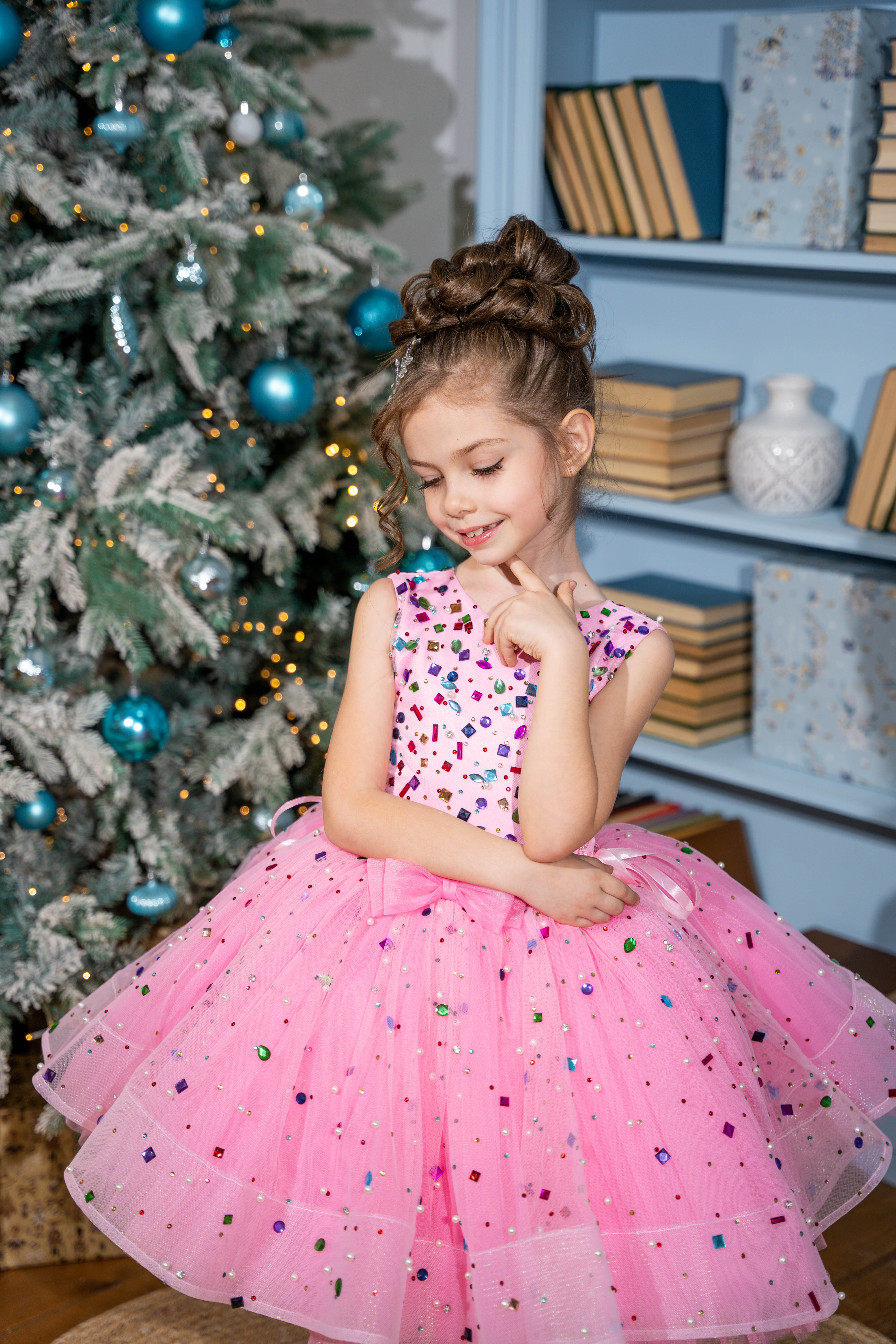 Baby girl in a pink dress with colorful sequins standing in front of a decorated Christmas tree.
