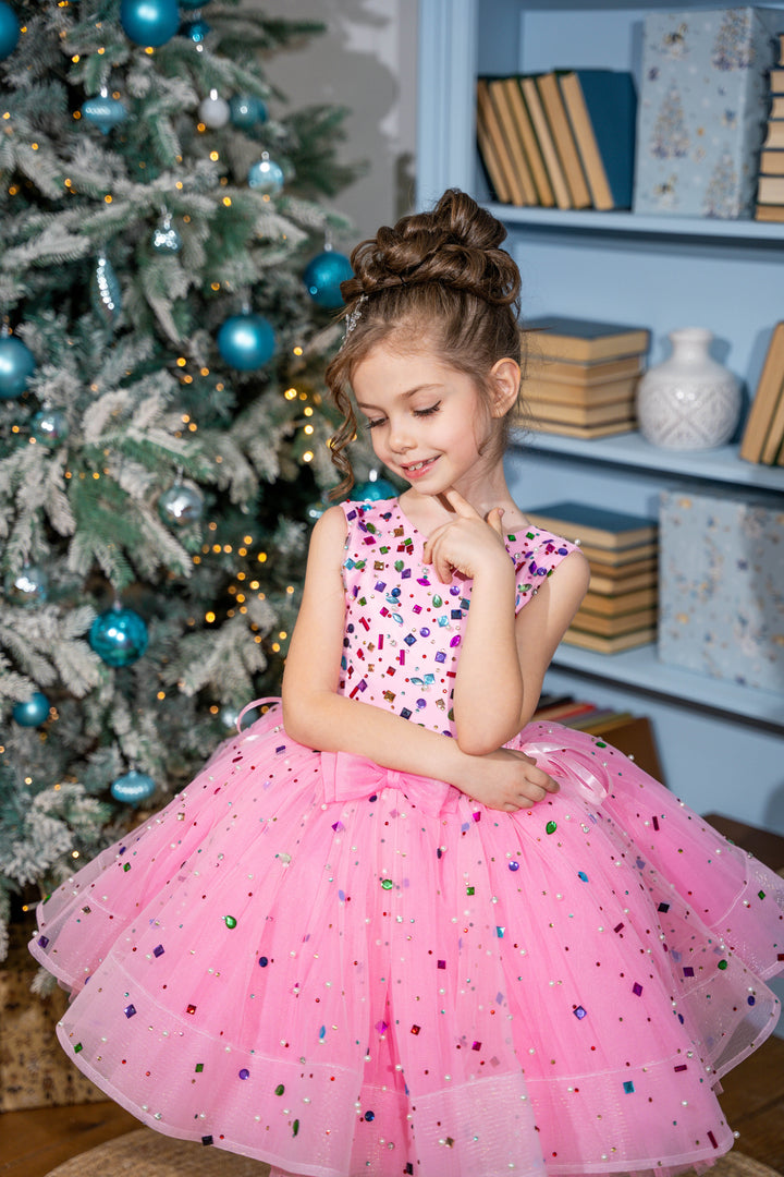 Baby girl in a pink dress with colorful sequins standing in front of a decorated Christmas tree.