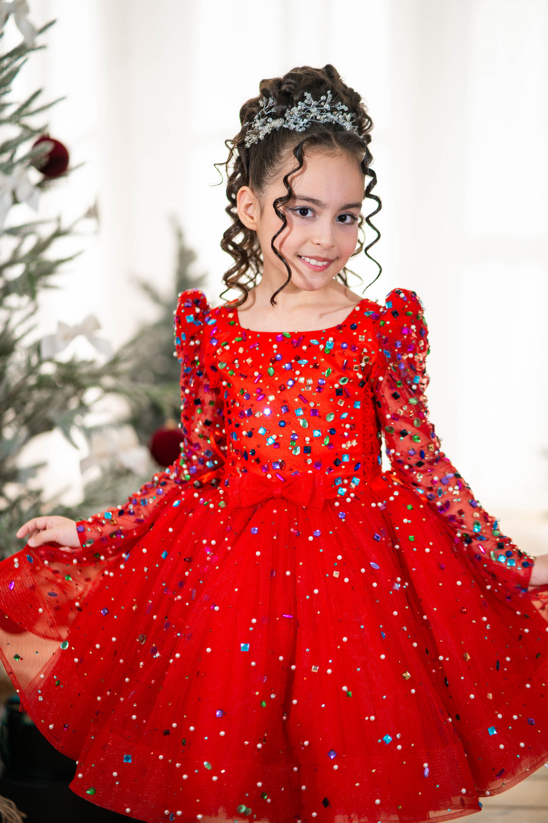 Baby girl in a red sequined dress standing in front of a Christmas tree.