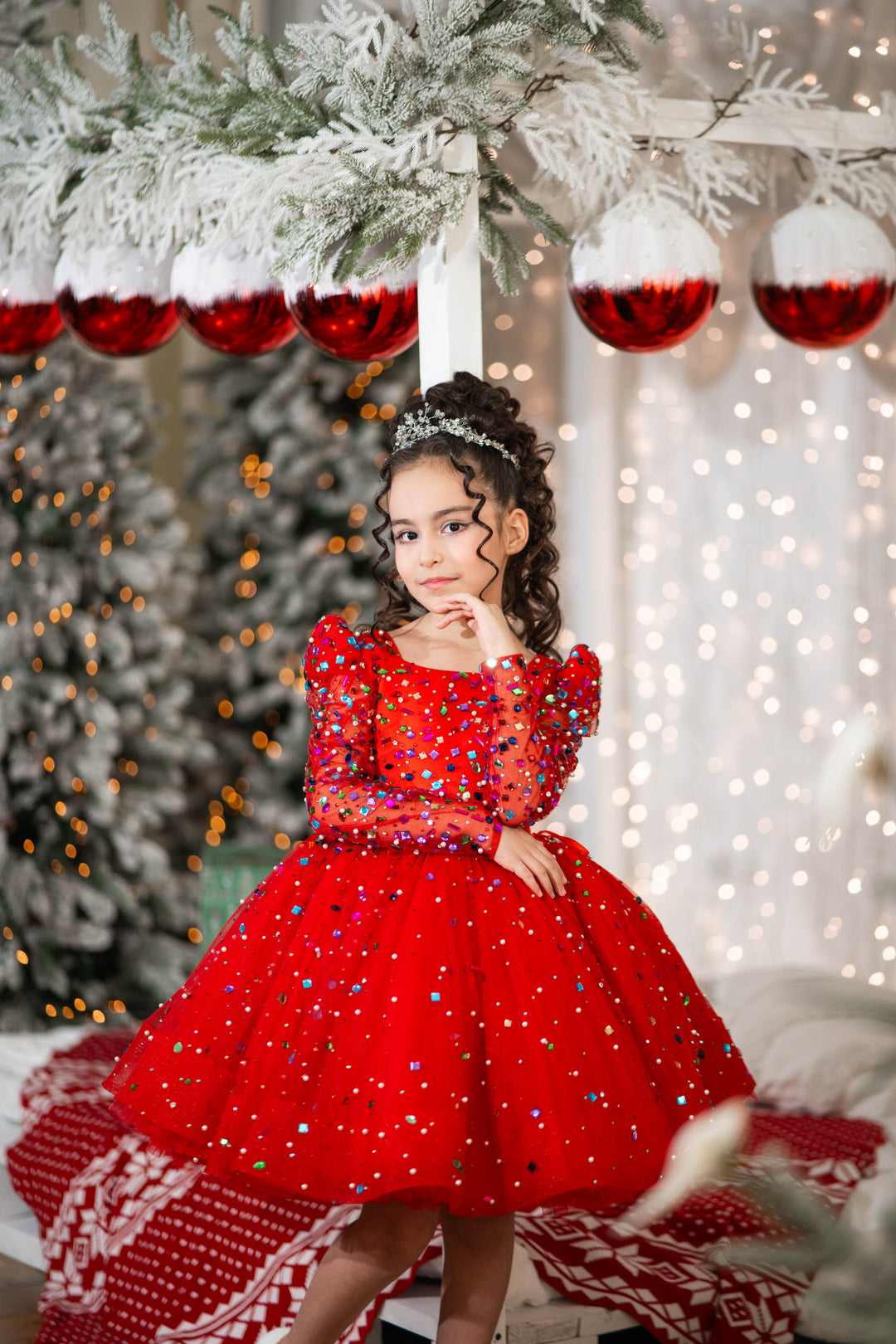 Young girl in a red dress standing in front of a decorated Christmas tree with red ornaments.