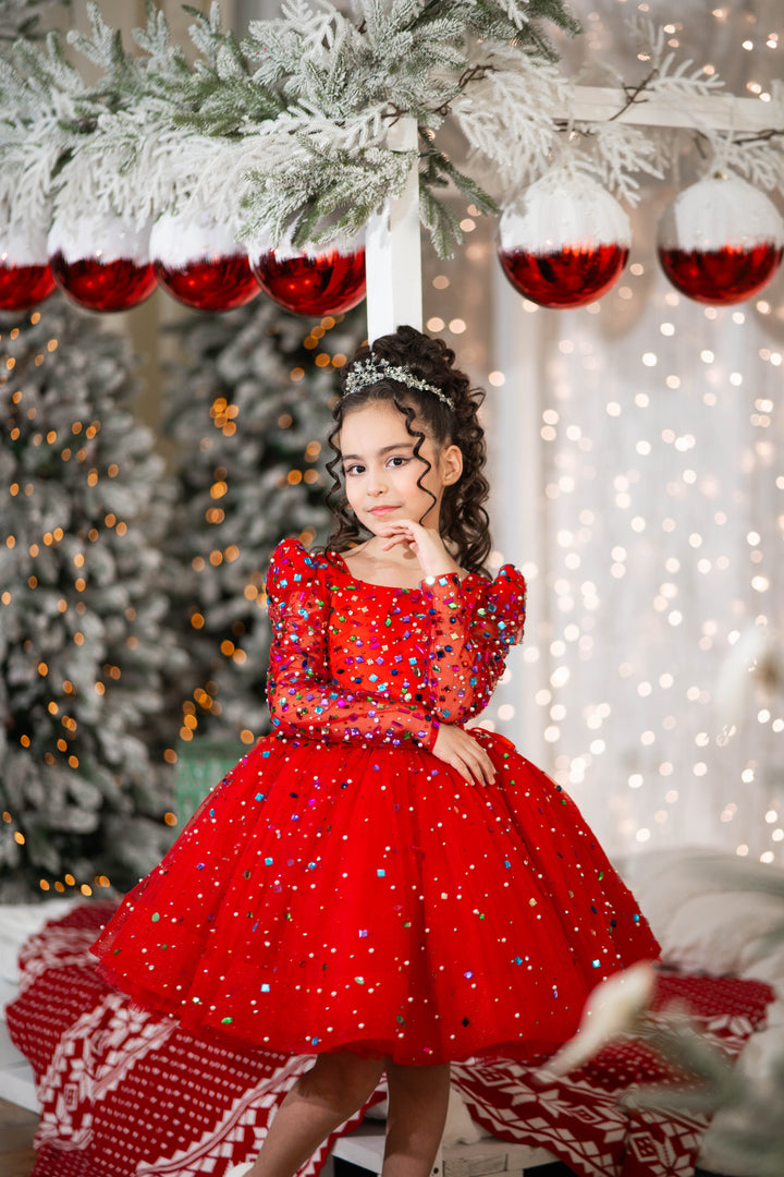 Young girl in a red dress standing in front of a decorated Christmas tree with red ornaments.