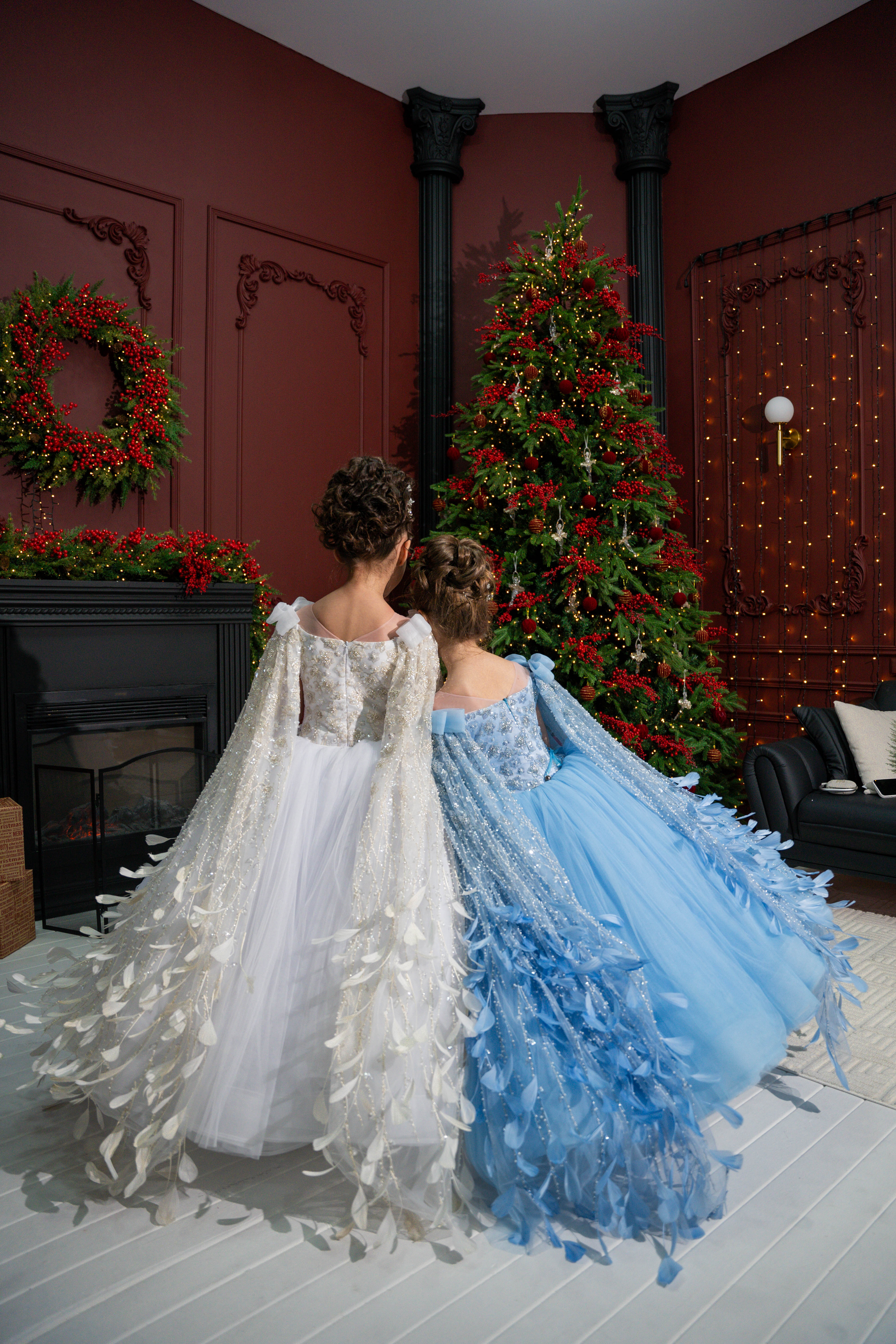 Two children in white and blue dresses standing in a room with a Christmas tree and wreath.