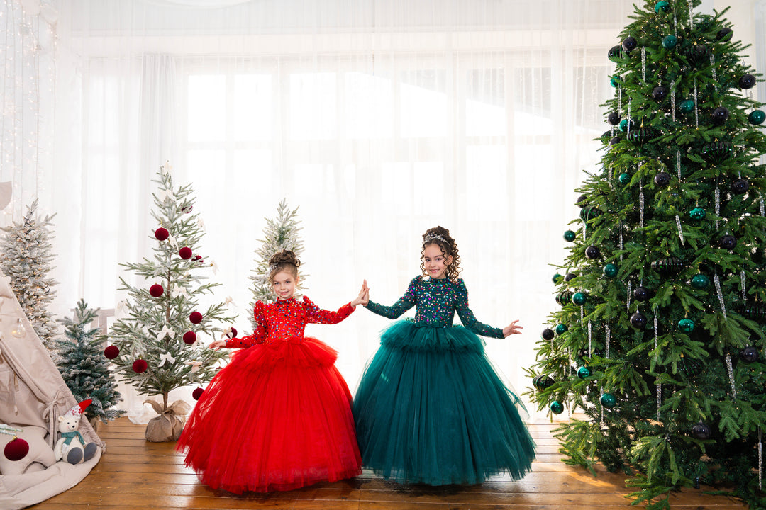 Two children in red and teal dresses standing in front of Christmas trees.