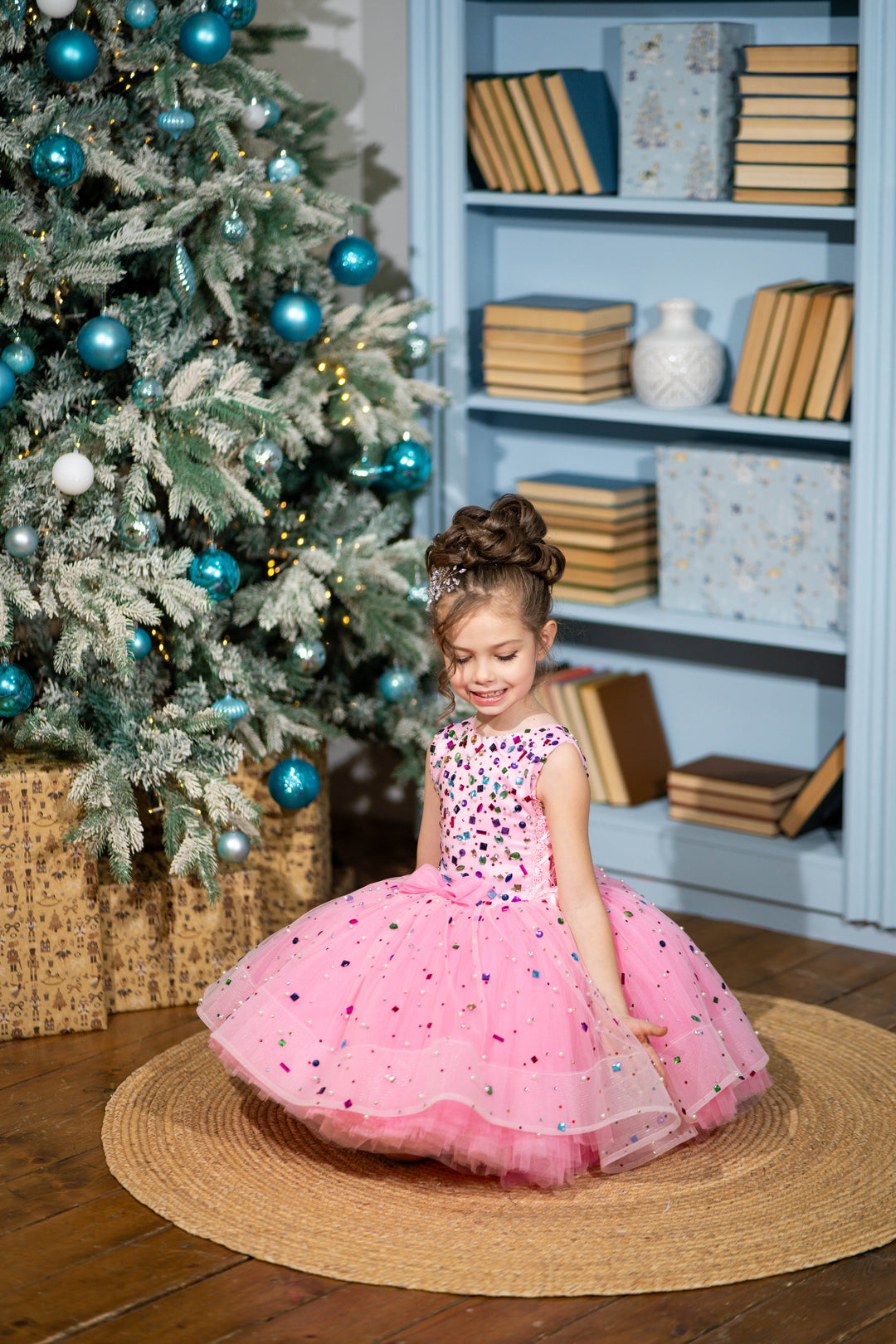 Baby girl in a pink dress sitting on the floor next to a decorated Christmas tree.
