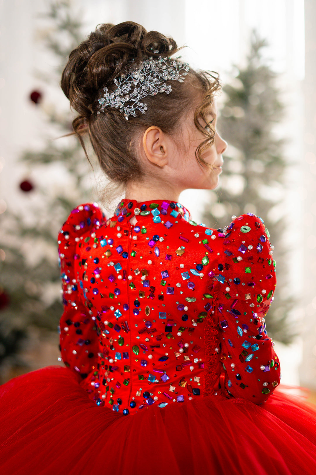 Child in a red dress with sequins and a headpiece, standing in front of a blurred festive background.