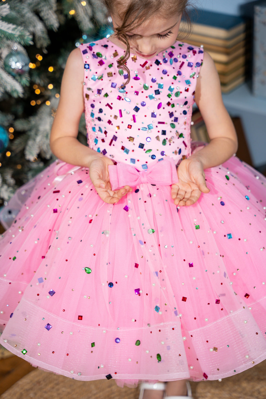 Baby girl in a pink dress with colorful sequins in front of a decorated Christmas tree.