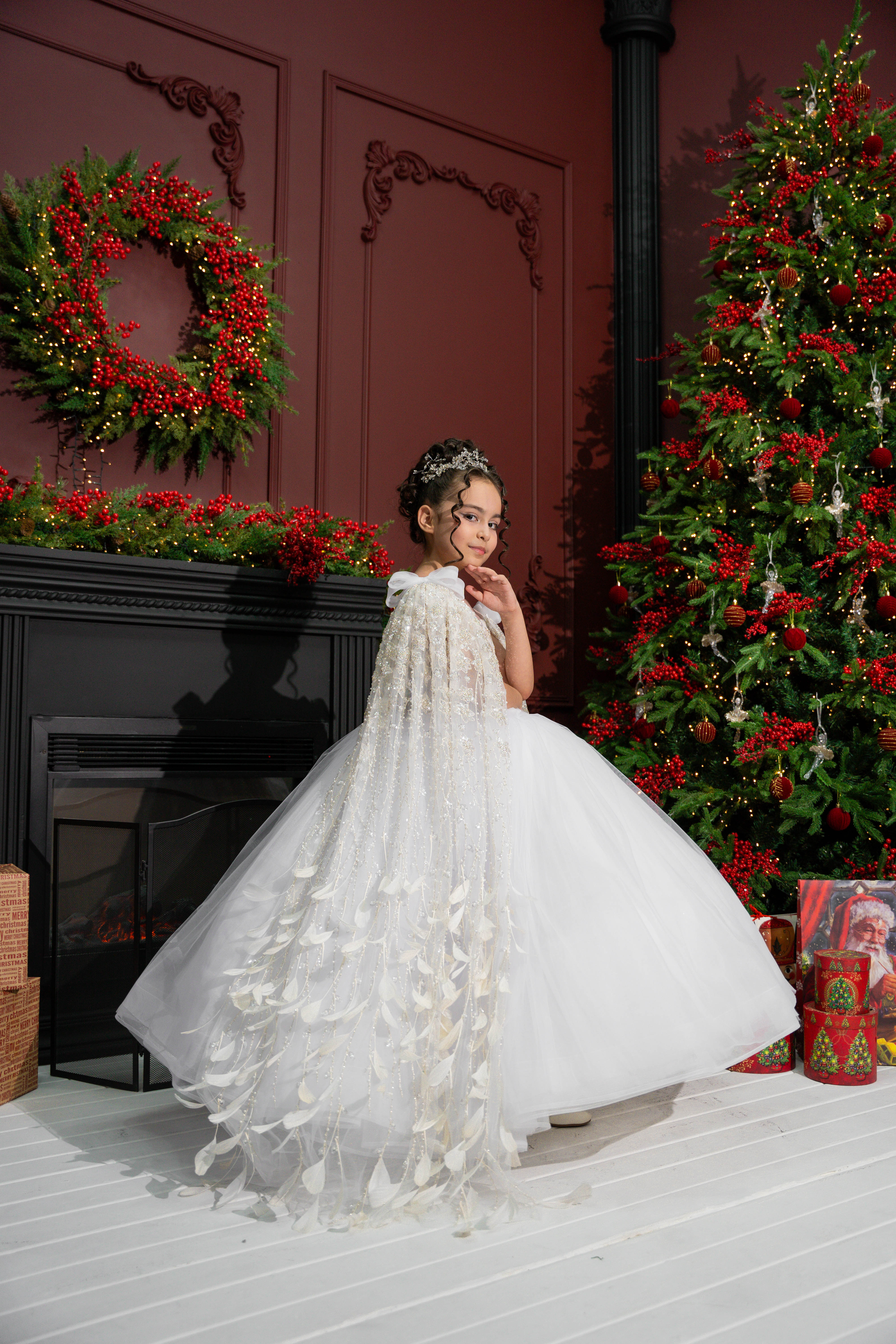 A child in a white dress standing in front of a decorated Christmas tree and fireplace.