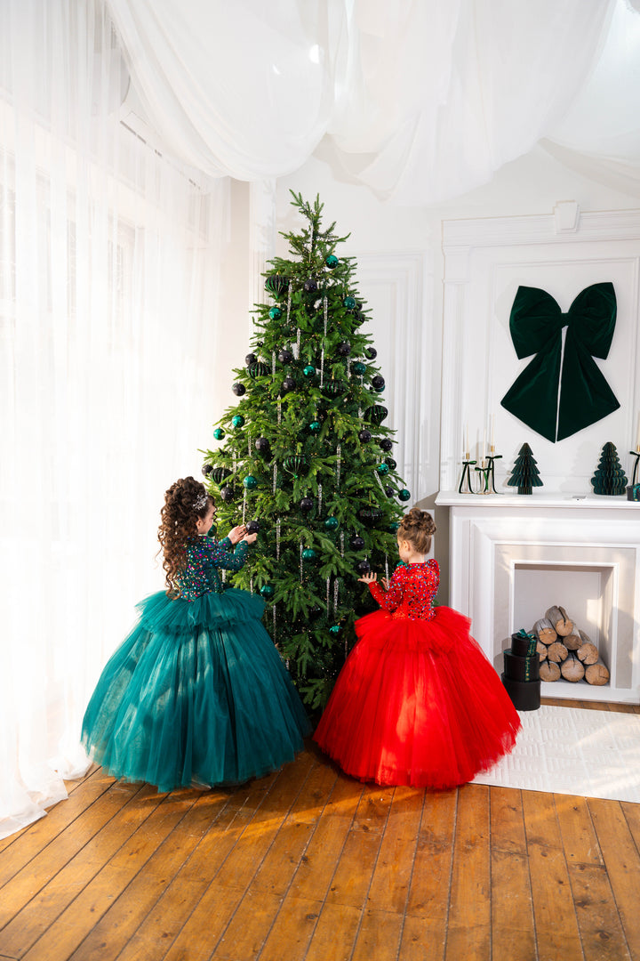 Two children in colorful dresses standing next to a decorated Christmas tree in a room with wooden floors and white walls.